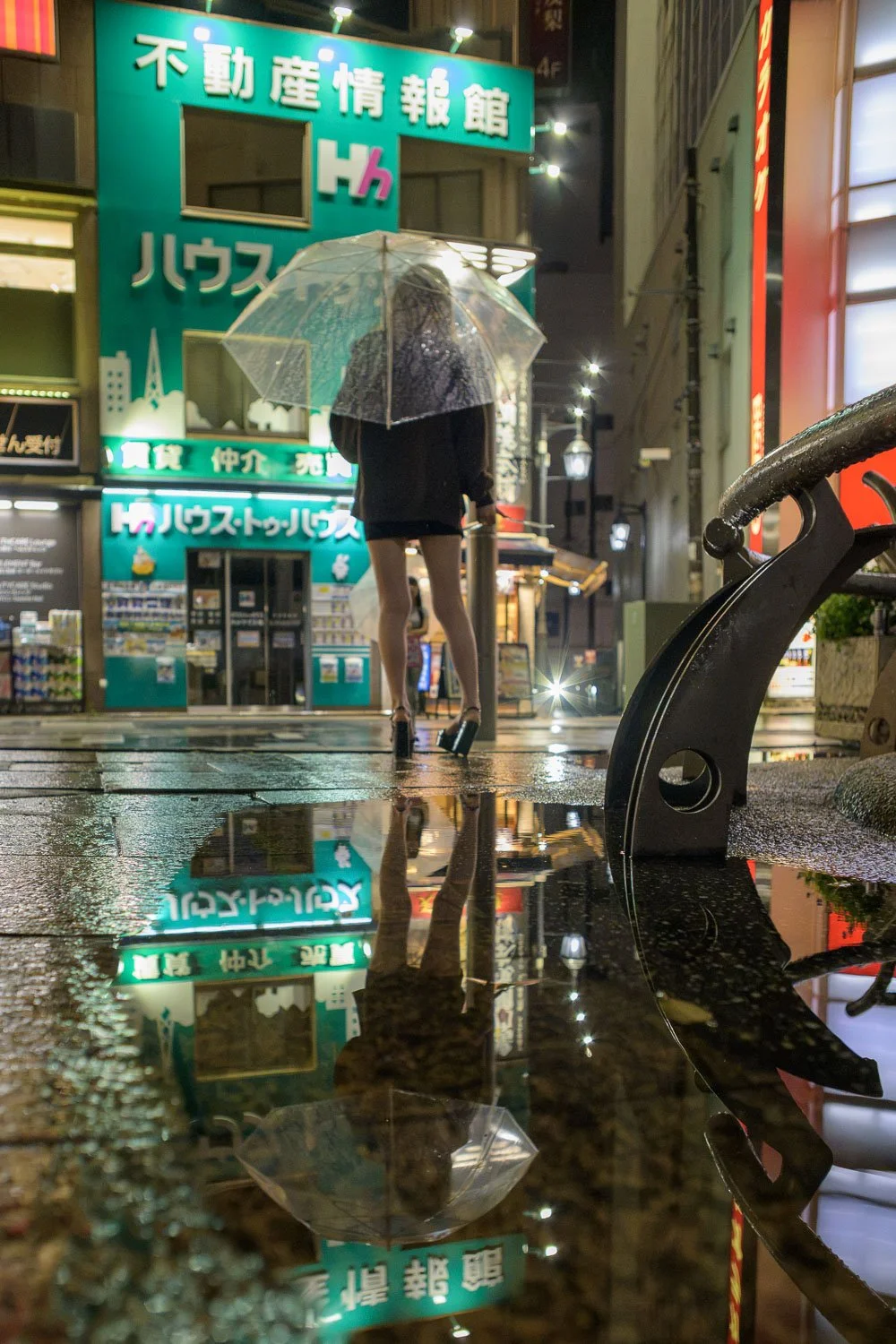 A woman under a clear umbrella reflected in a rain puddle beneath green neon signage, Akabane at night