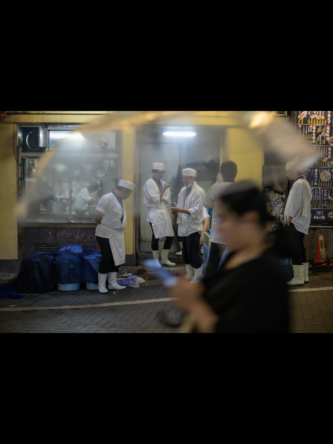Cooks in white caps and rubber boots take a break outside an izakaya kitchen in Shibuya at night, a passer-by blurred in the foreground, forged at 50mm on a Nikon Z9