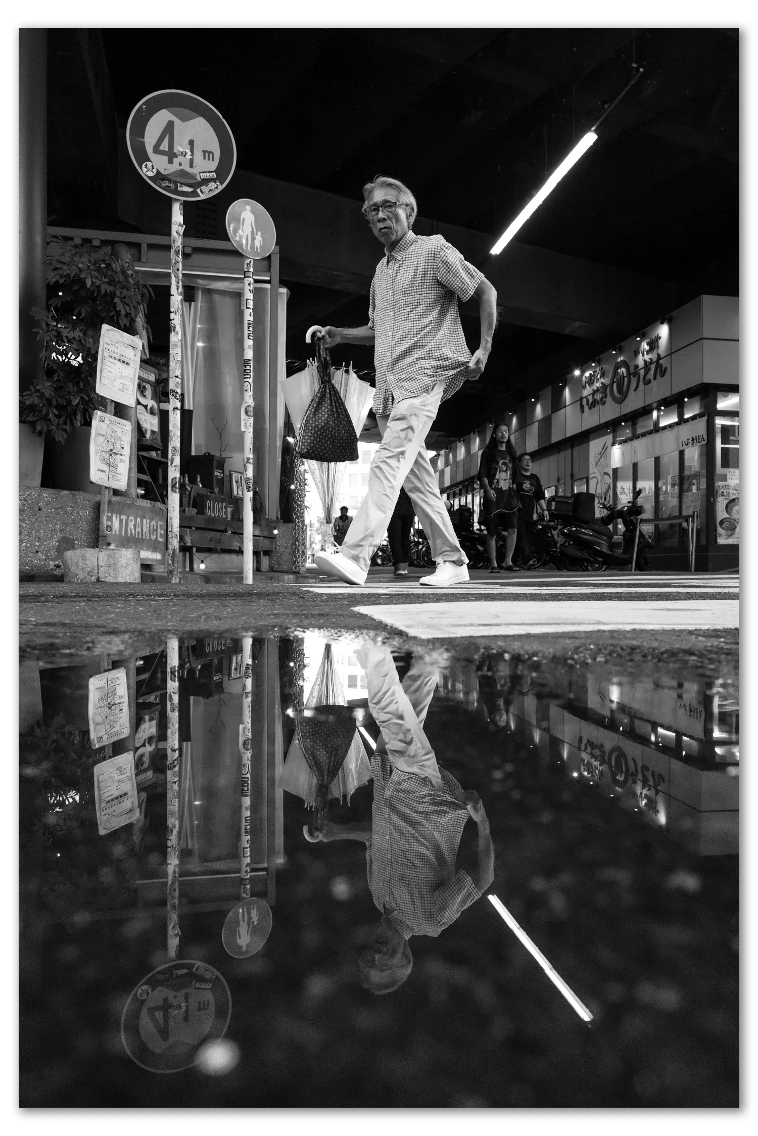 Black and white street photograph, Nakano, Tokyo — a man crosses an arcade entrance mirrored in a rain puddle, Leica Q2 Monochrom 28mm