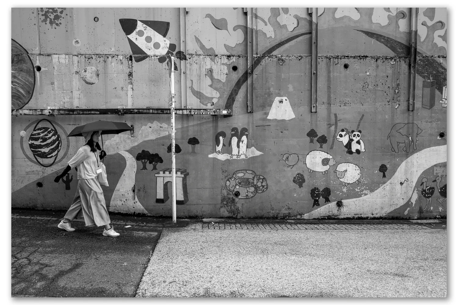 Black and white street photograph, Nakano, Tokyo — a woman with an umbrella passes a concrete floodwall painted with penguins, pandas and Mount Fuji, Leica Q2 Monochrom 28mm