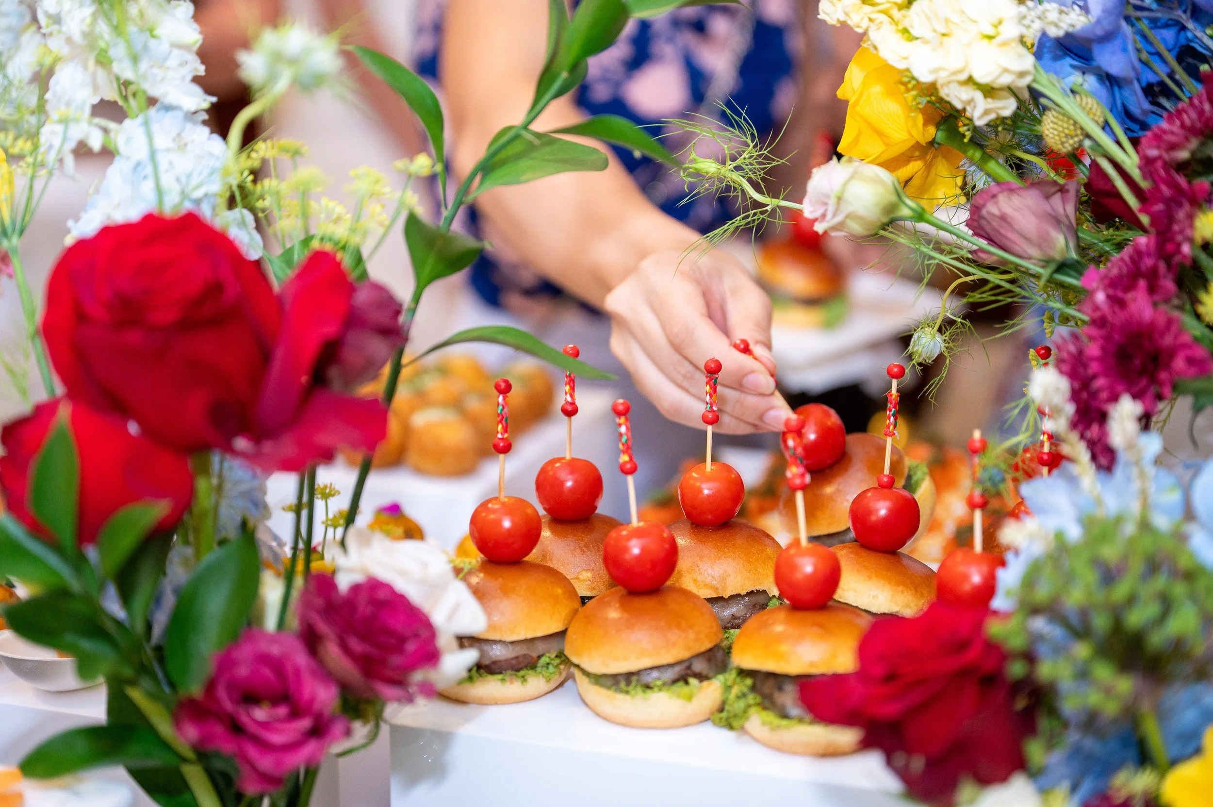 A person’s hand decorating small sliders with cherry tomatoes on top, surrounded by colorful flowers and greenery.