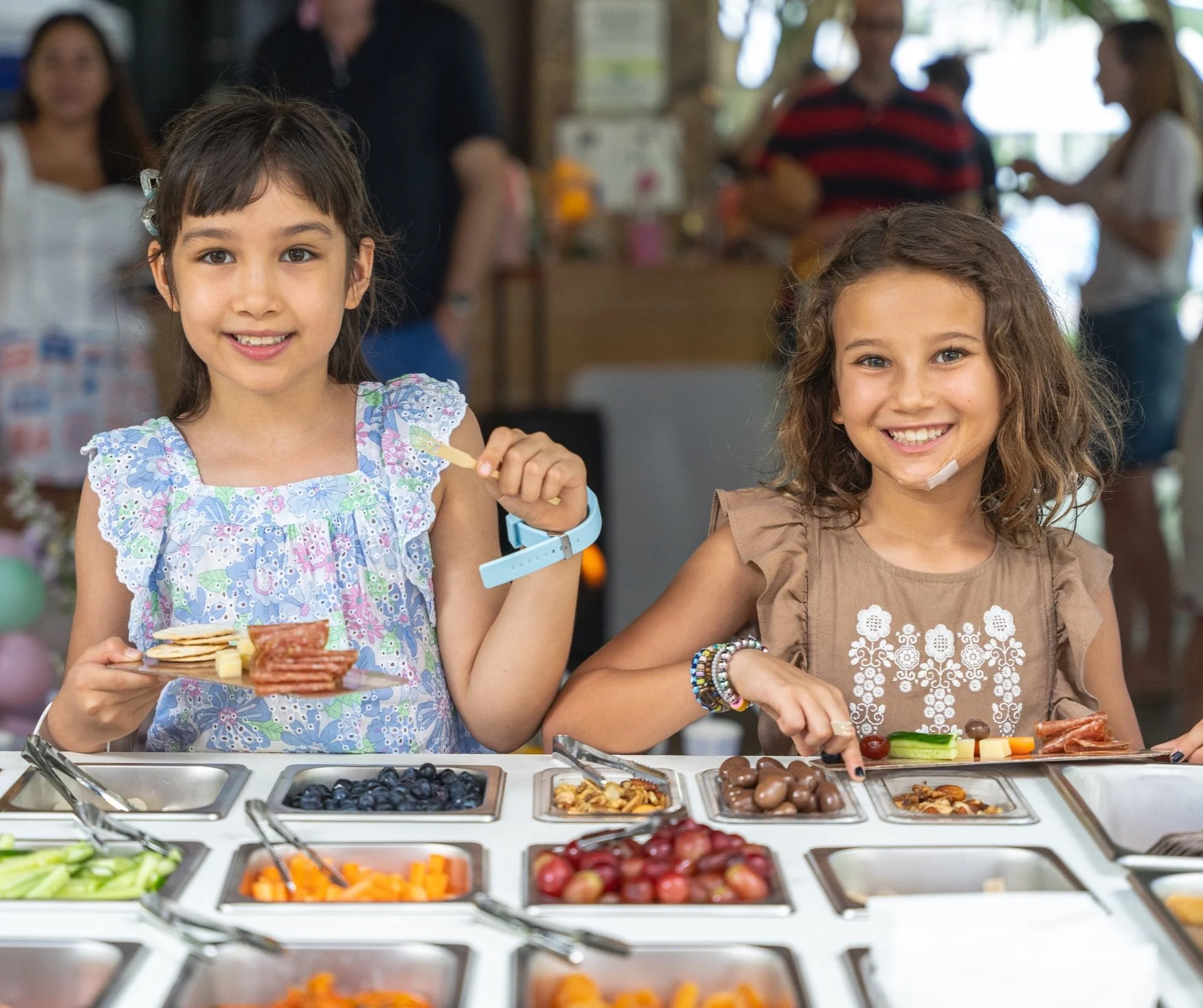 Two young girls smiling at a buffet with various fruits and snacks, enjoying a snack at an indoor celebration.