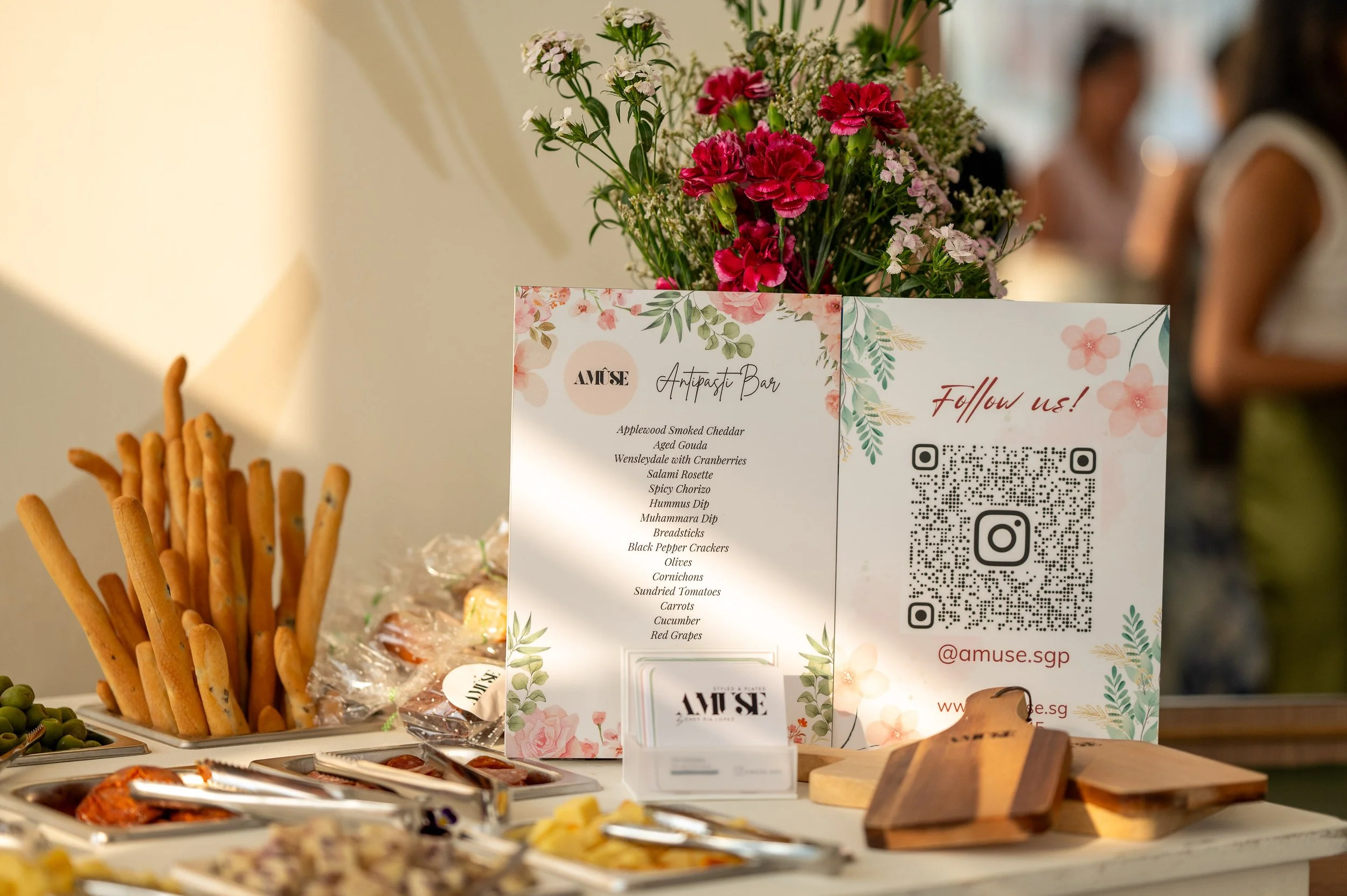 A floral signboard for an appetizer bar with a menu, QR code, and social media handle, surrounded by breadsticks, olives, and other snacks, with a bouquet of pink and white flowers in the background.