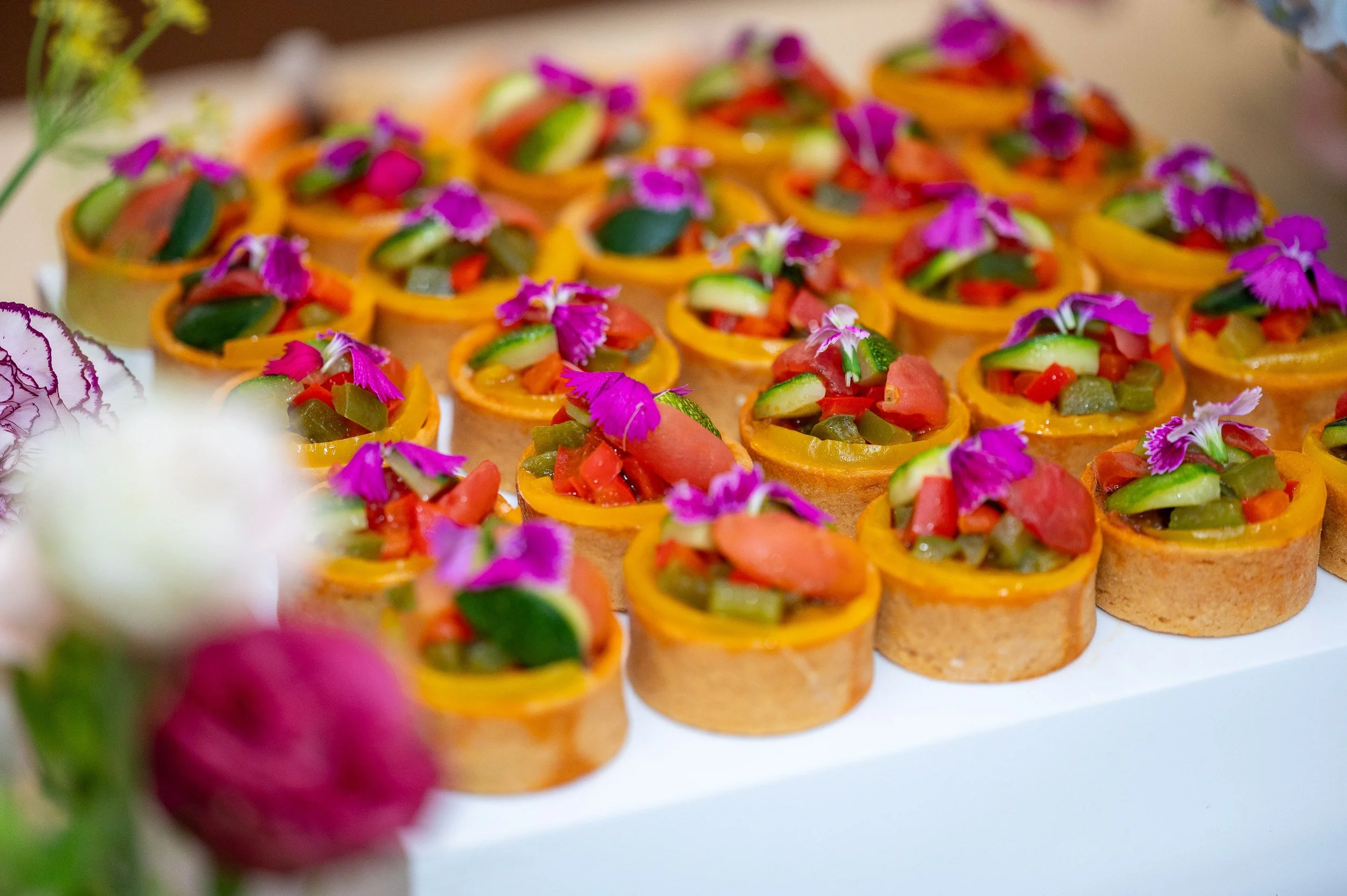Tray of bite-sized appetizers topped with vegetables and purple edible flowers.