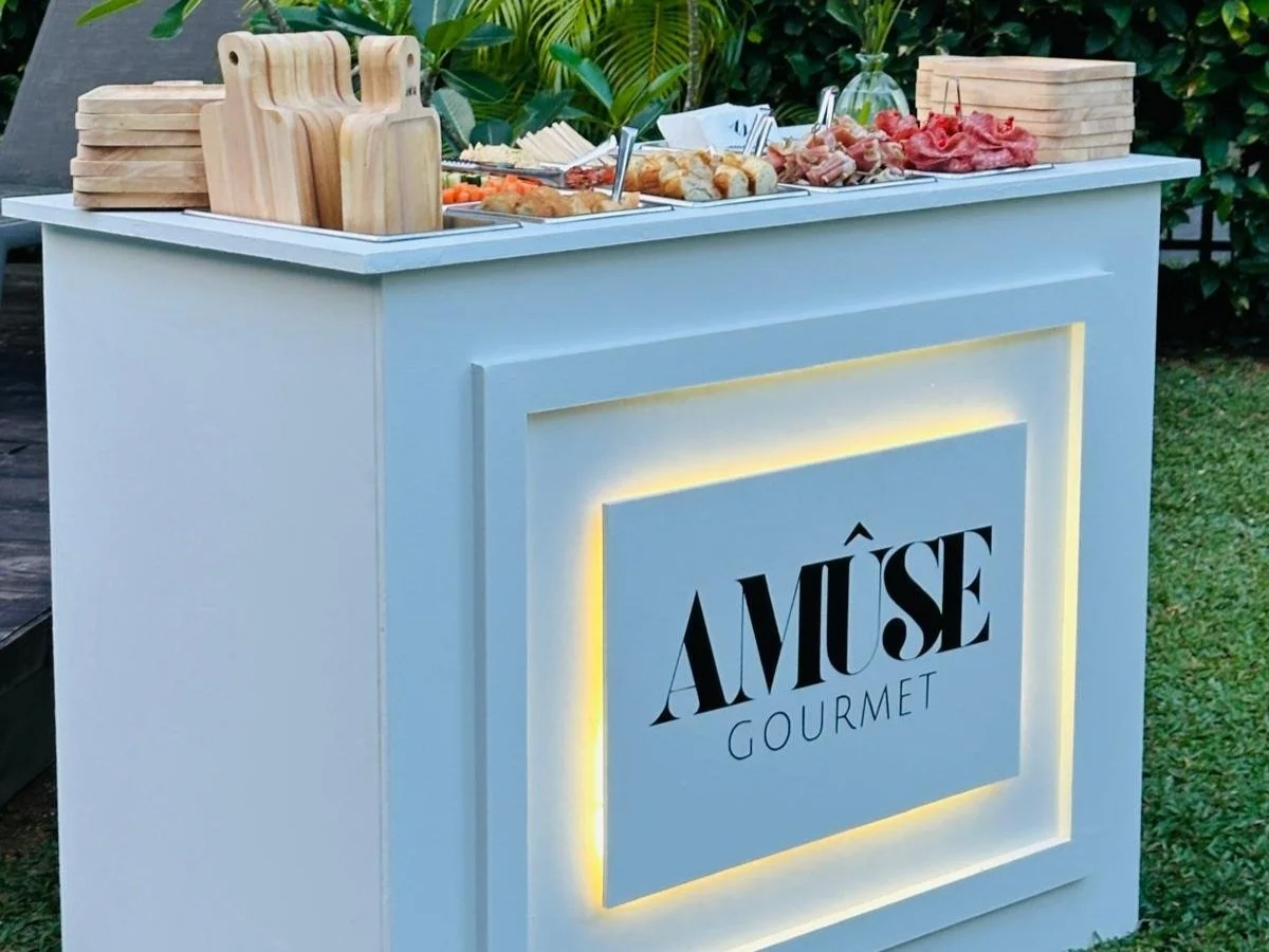 Outdoor buffet table with wooden cutting boards, cold cuts, bread, and vegetables, in front of a sign that reads 'AMÜSE GOURMET' illuminated from behind.