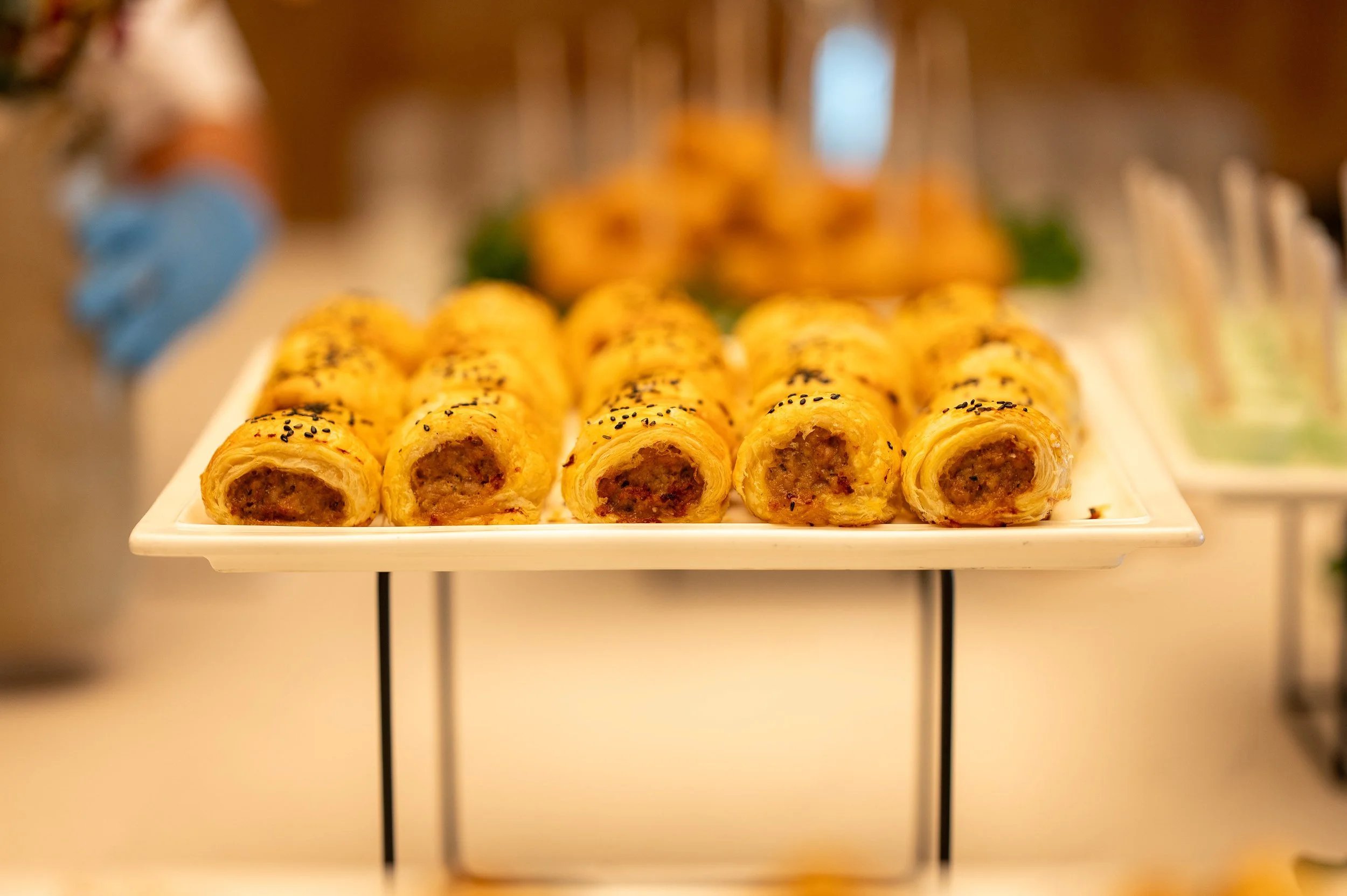 A white rectangular platter holding small sausage and cheese pastries with sesame seeds on top, displayed on a tiered stand at a buffet.
