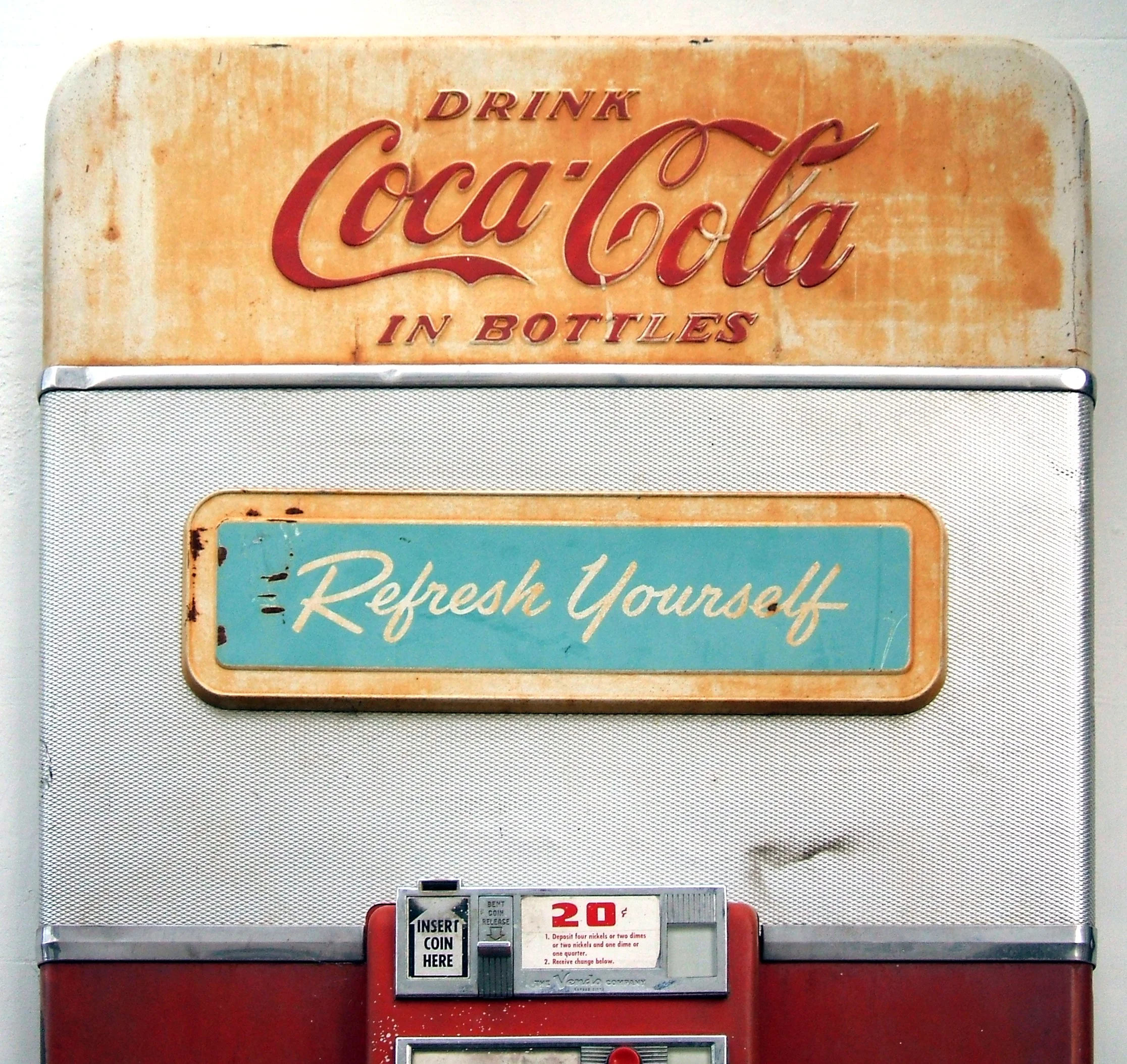 An old Coca-Cola vending machine in East Sussex, England. Along the top of the machine, it says 'Drink Coca-Cola in bottles', and underneath that, it says 'Refresh yourself'. A coin machine allows you to buy a bottle, which costs 20 cents.