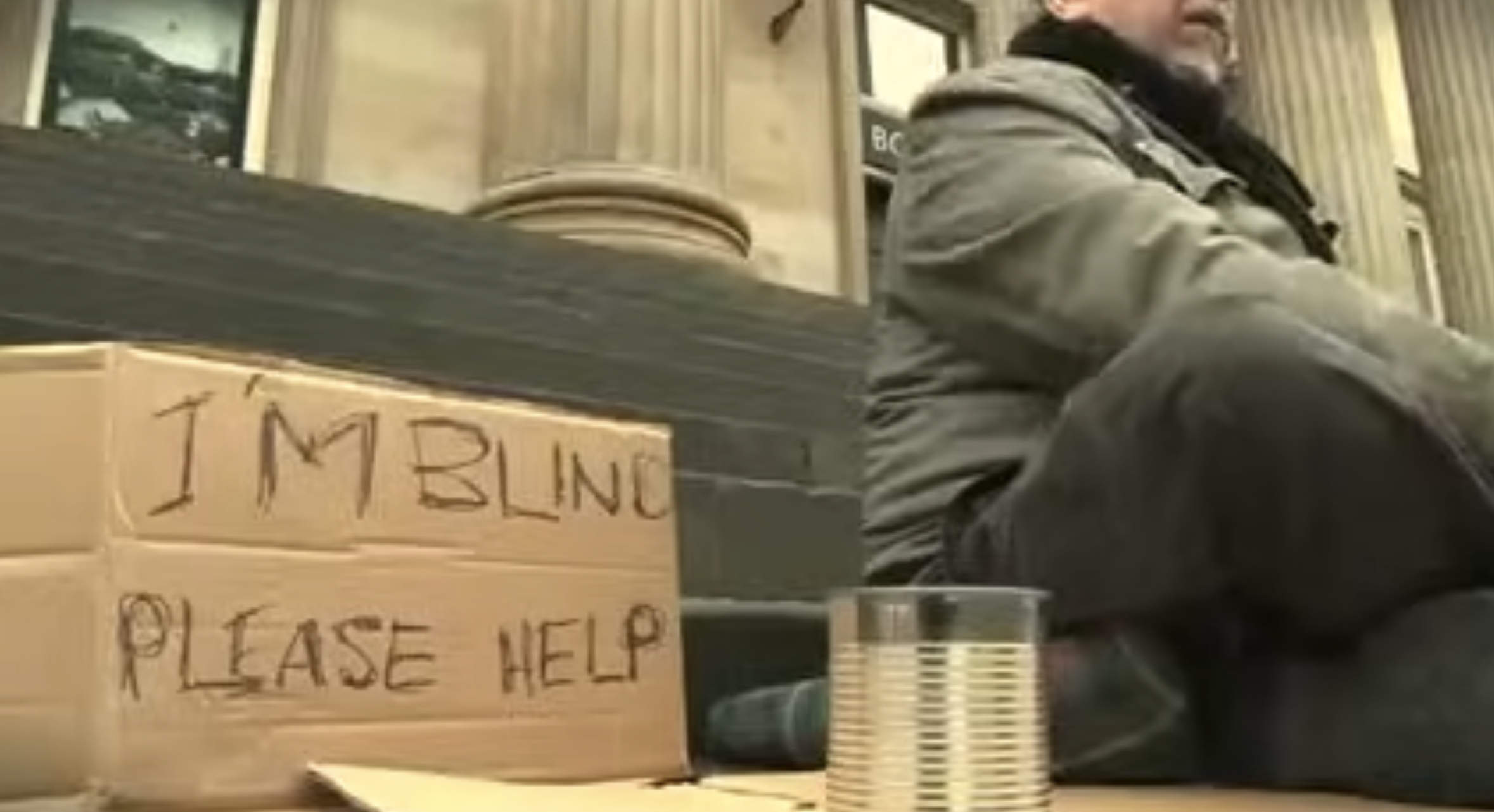 A man sits cross-legged in a public square with a sign beside him that reads, I'm blind, please help. There is an empty tin can in front of the sign.