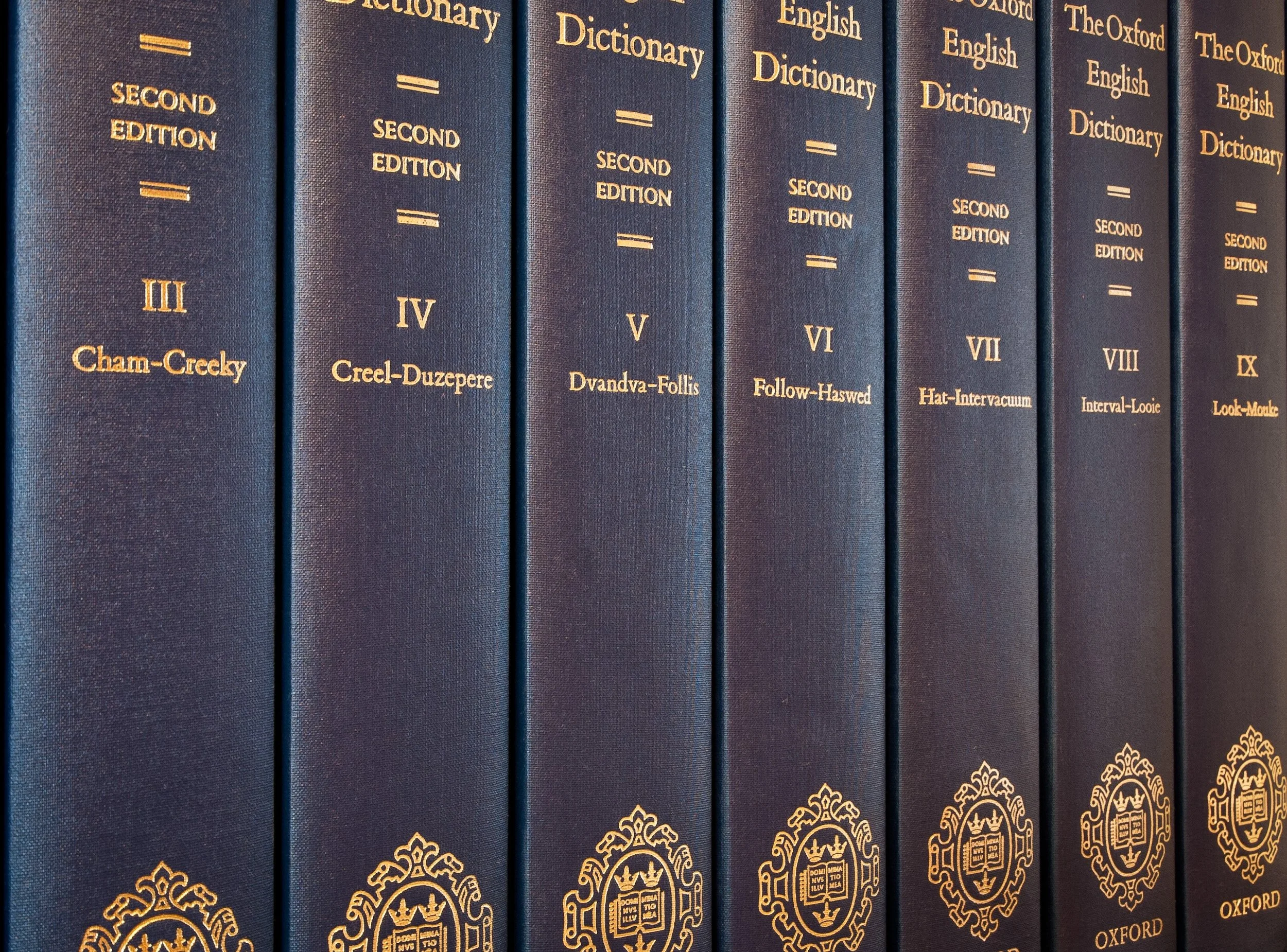 A close-up of 7 hardback copies of the Oxford English Dictionary, 2nd edition, lined up on a bookshelf.