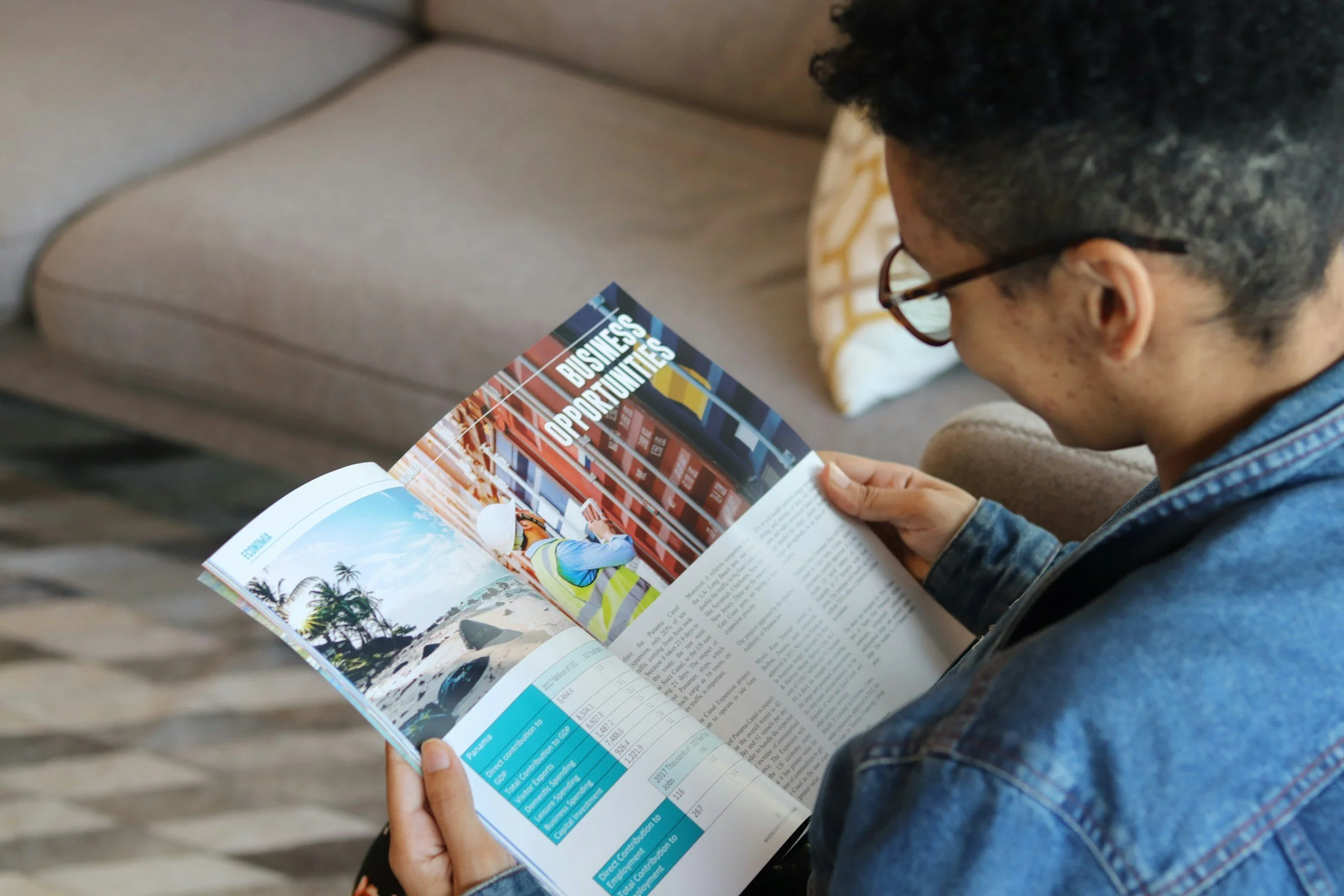 A man sits on a couch smiling while reading an economics magazine.