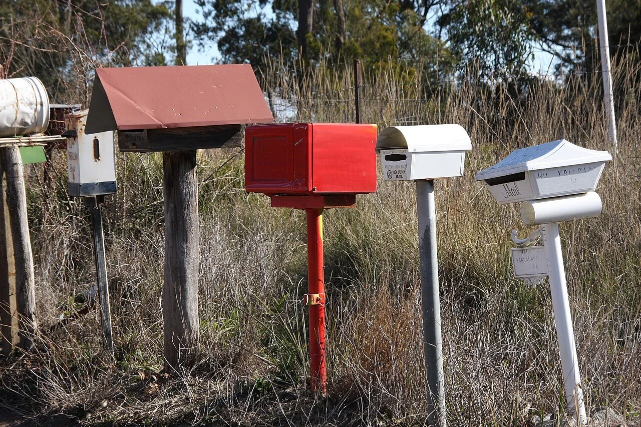 Letterboxes appear in a line one after the other, in a rural setting. They are all different shapes and sizes.