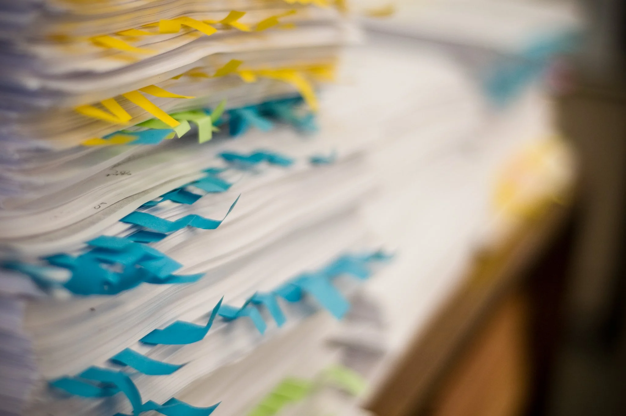 A tall pile of paper is stacked on top of a desk. Blue and yellow tags are sticking out from many of the pages in the pile.
