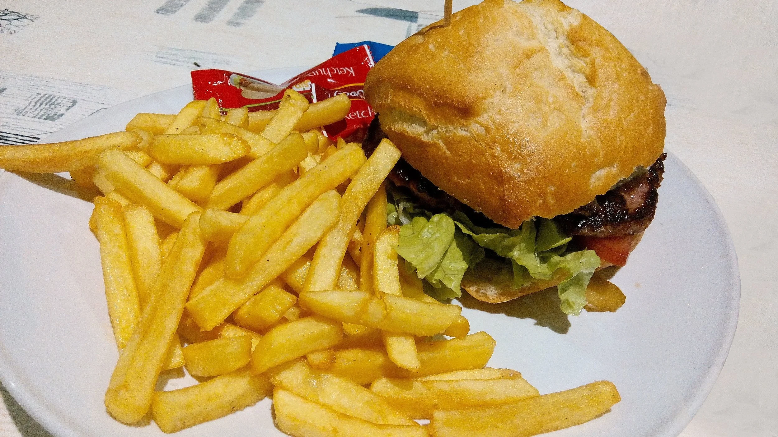 A white dinner plate has on it a burger and a pile of chips and a crumpled up sachet of tomato sauce.