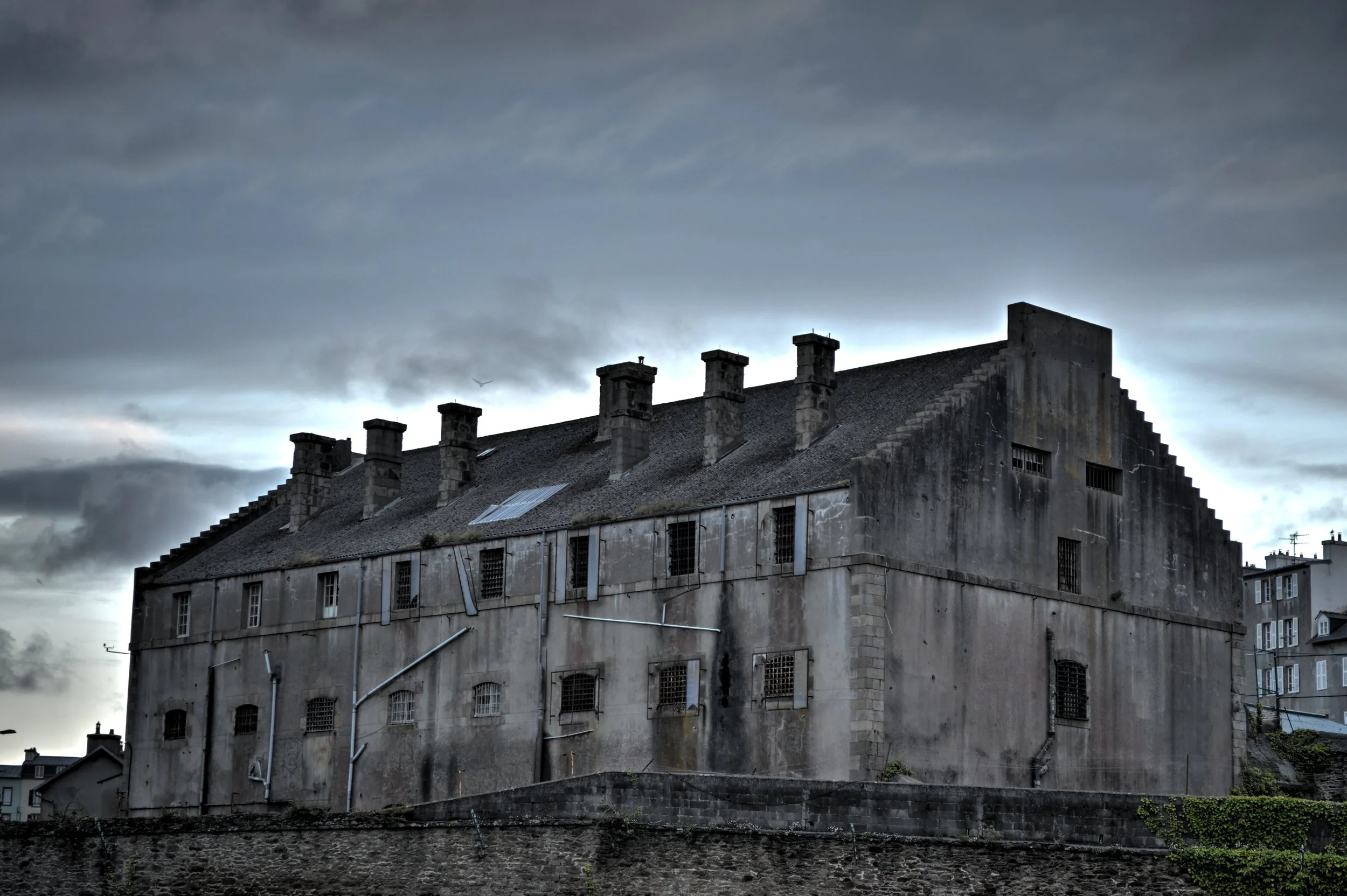 Old abandoned prison building with barred windows under a dark moody sky.