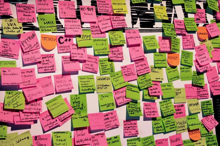 A white wall is almost entirely covered in pink and green, and some yellow and orange, post-it notes. Each note contains black writing that takes up most of the space on the note.
