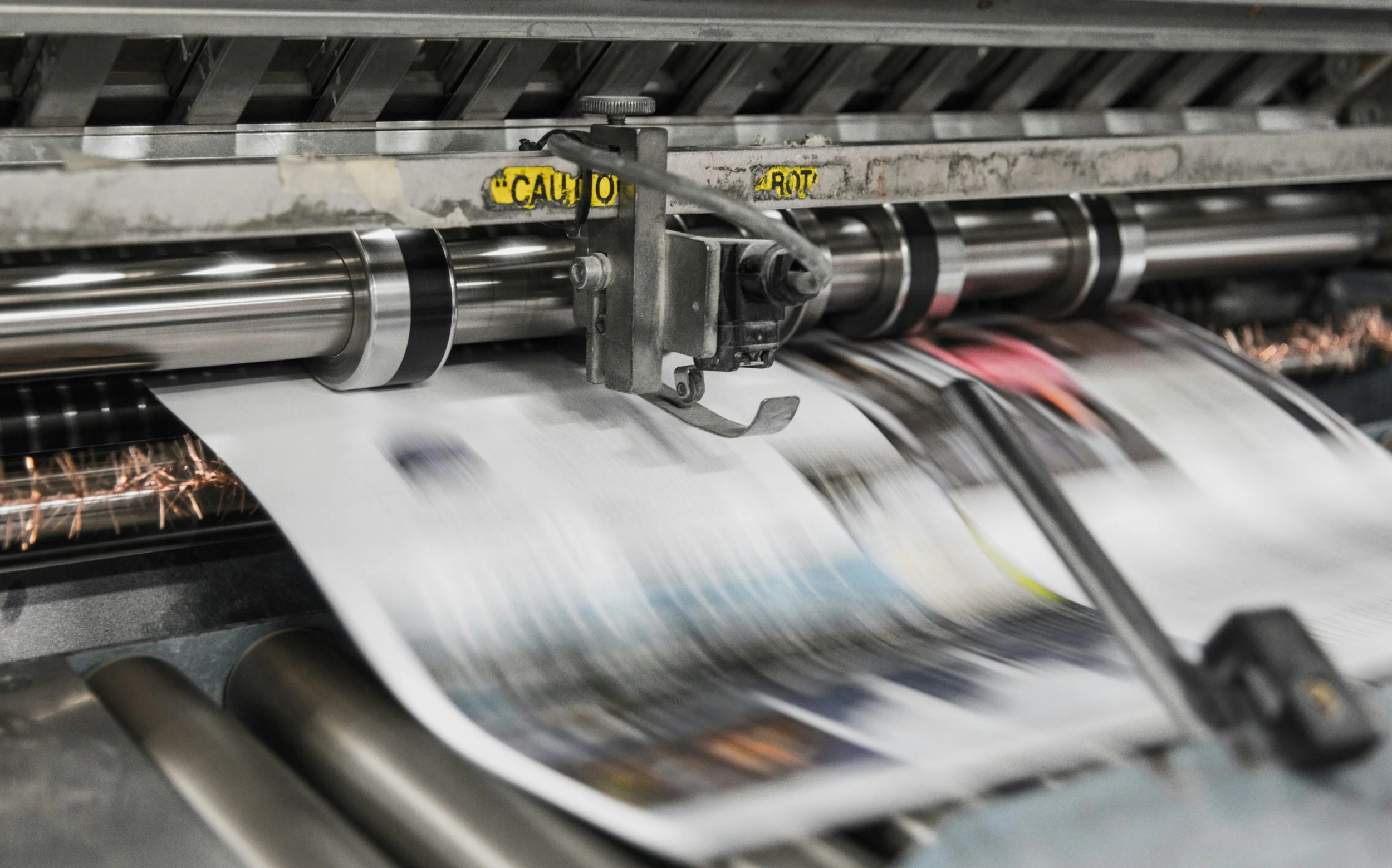Pages come off a printing press. The pages' contents are blurry because of how fast they come out of the press.