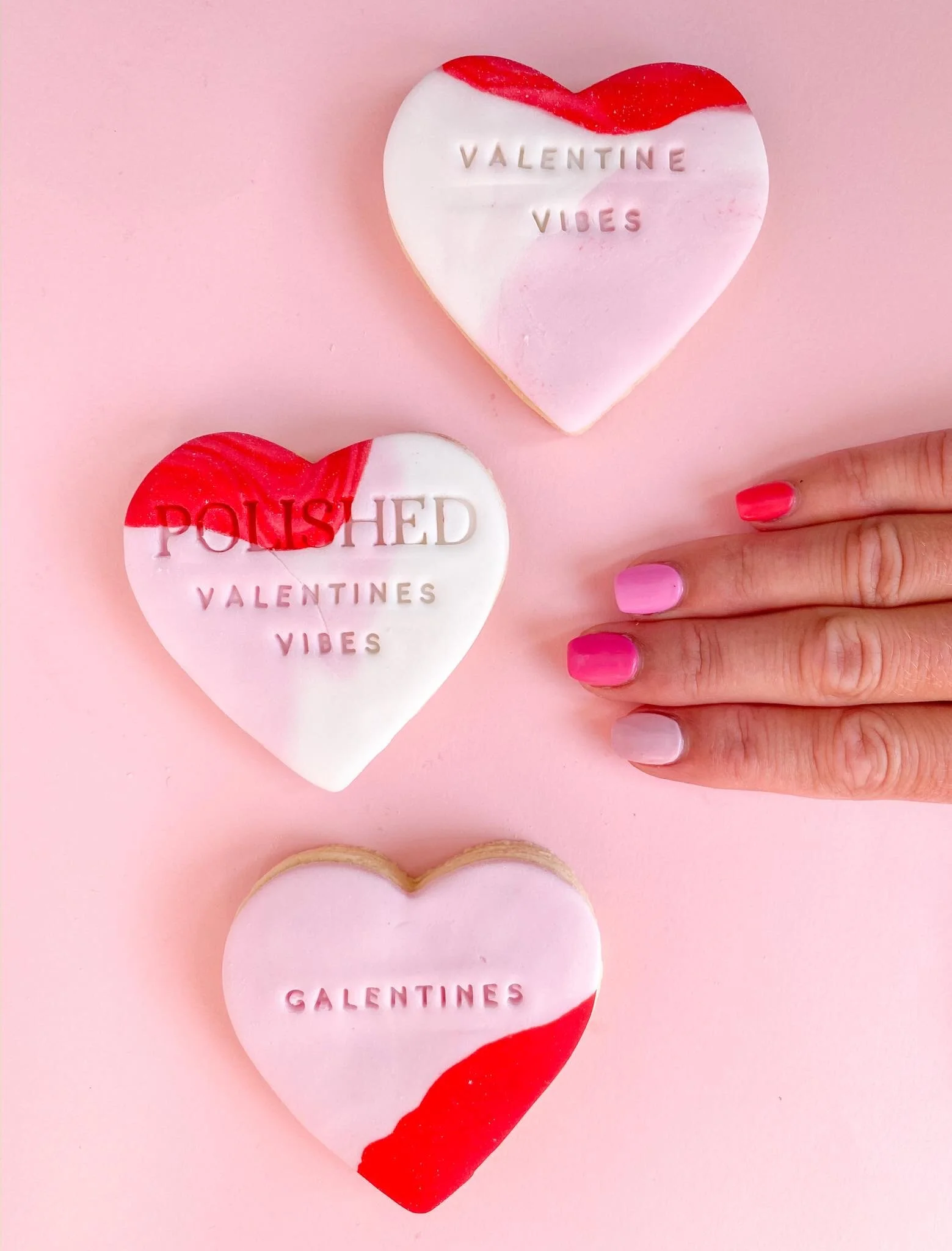 Valentine-themed heart-shaped soaps with red and white colors, inscriptions reading 'Valentine Vibes,' 'Poished Valentines,' and 'Galentines,' placed on a pink background, with fingers displaying pink and white nail polish.