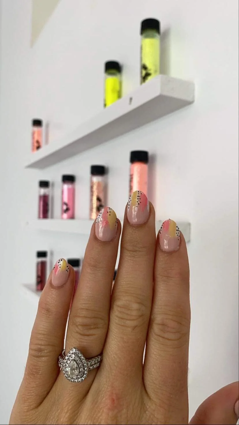 Close-up of a hand with a diamond ring and decorated nails with pink, yellow, and black dotted design. In the background, a white wall with three white shelves holding small bottles of colorful powders or liquids in various shades of pink, yellow, an