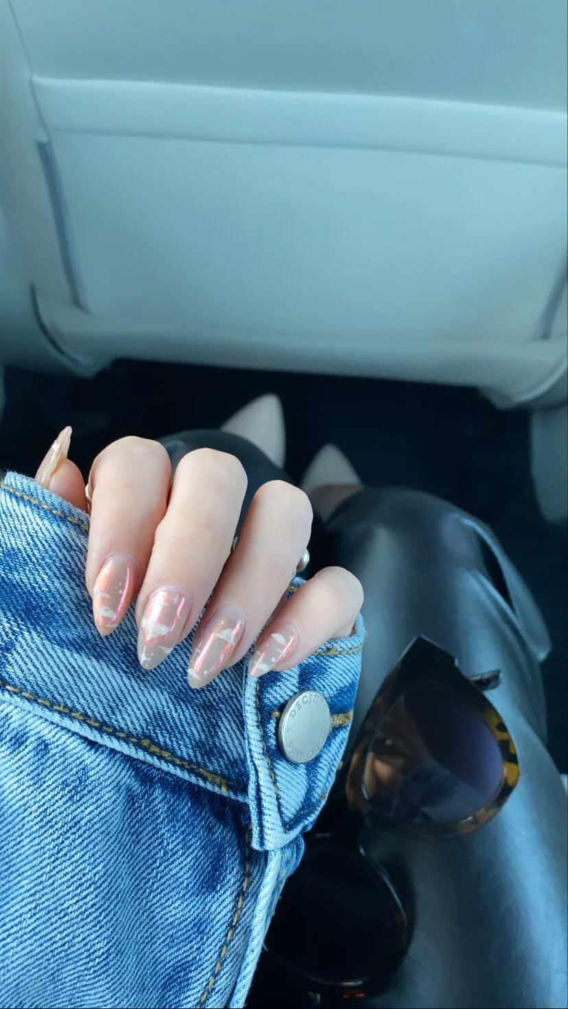 Person with painted nails resting their hand on the sleeve of a denim jacket, with sunglasses nearby, inside a vehicle.