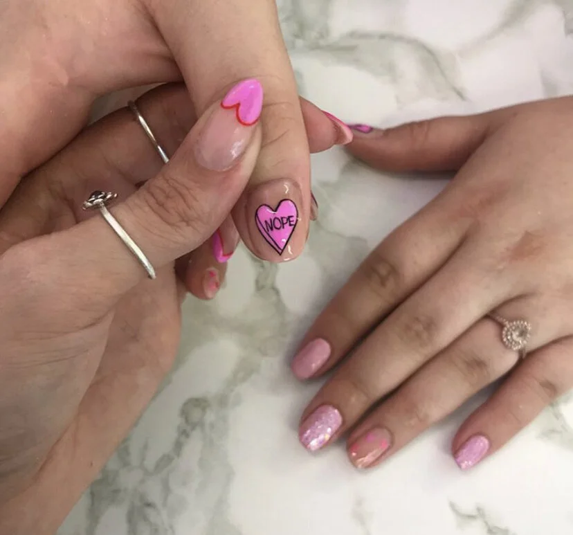 Close-up of two hands with pink heart-themed nail art, one hand showing a nail with the word 'NOPE' inside a pink heart, and the other with pink glittery nail polish, on a white marble surface.