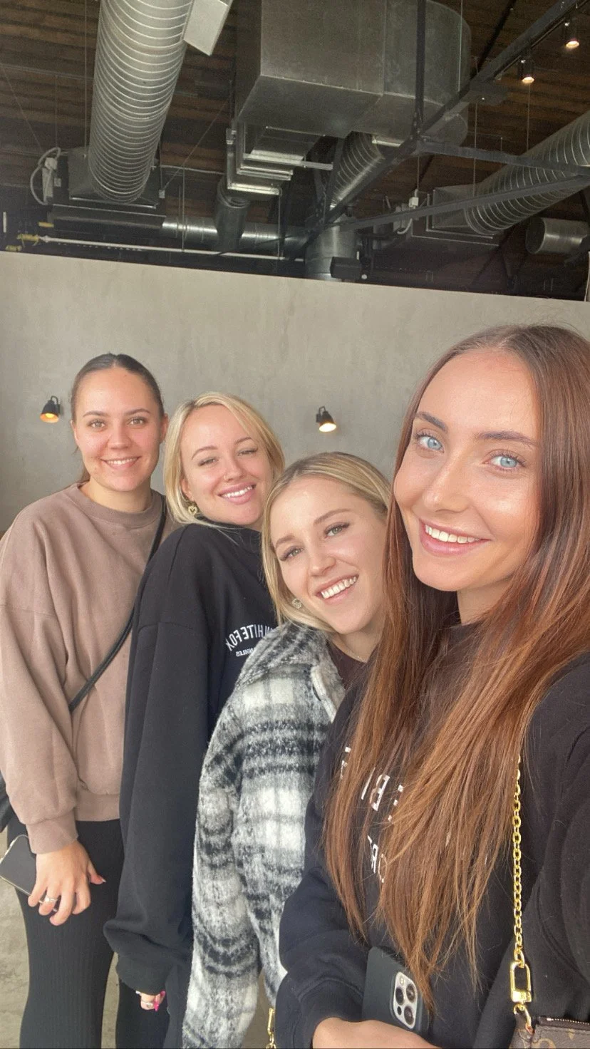 Four women smiling and taking a selfie in an industrial-style indoor space with visible ductwork on the ceiling.