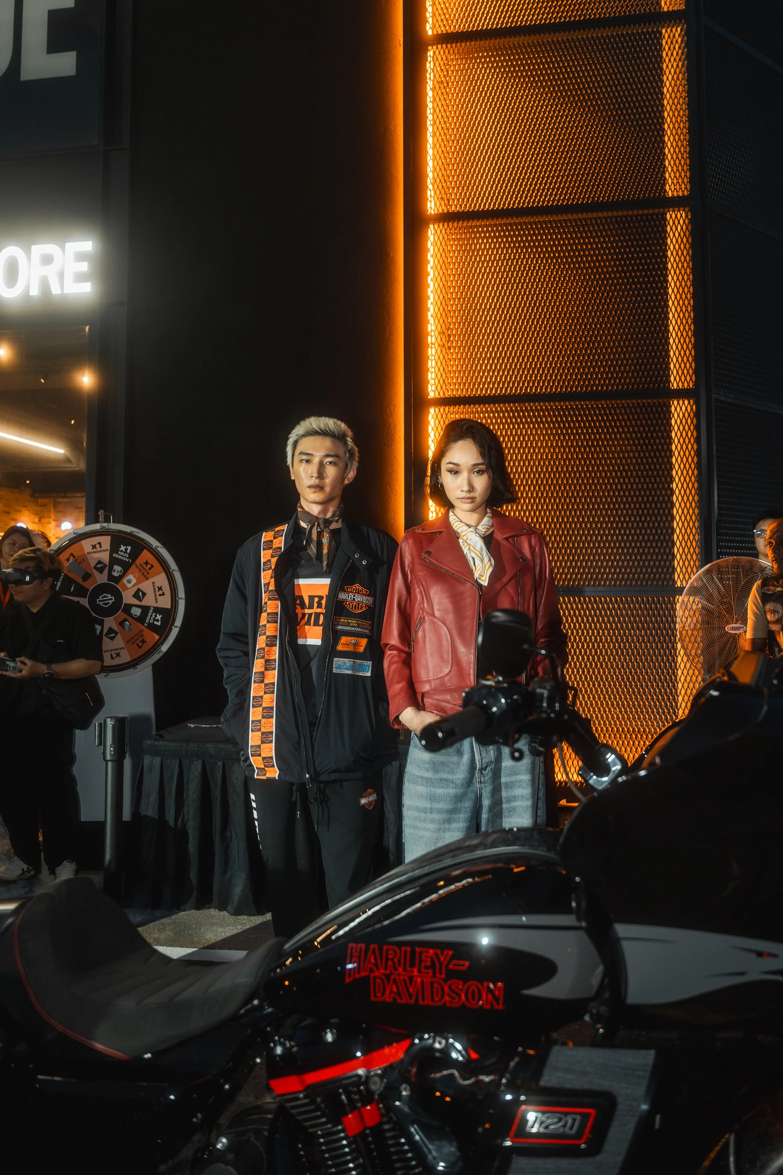 Two models standing next to a Harley-Davidson motorcycle at a nighttime event. The male model wears a black Harley-Davidson jacket with patches, and the female model wears a red leather jacket. A orange-lit panel and a crowd are visible in the background.