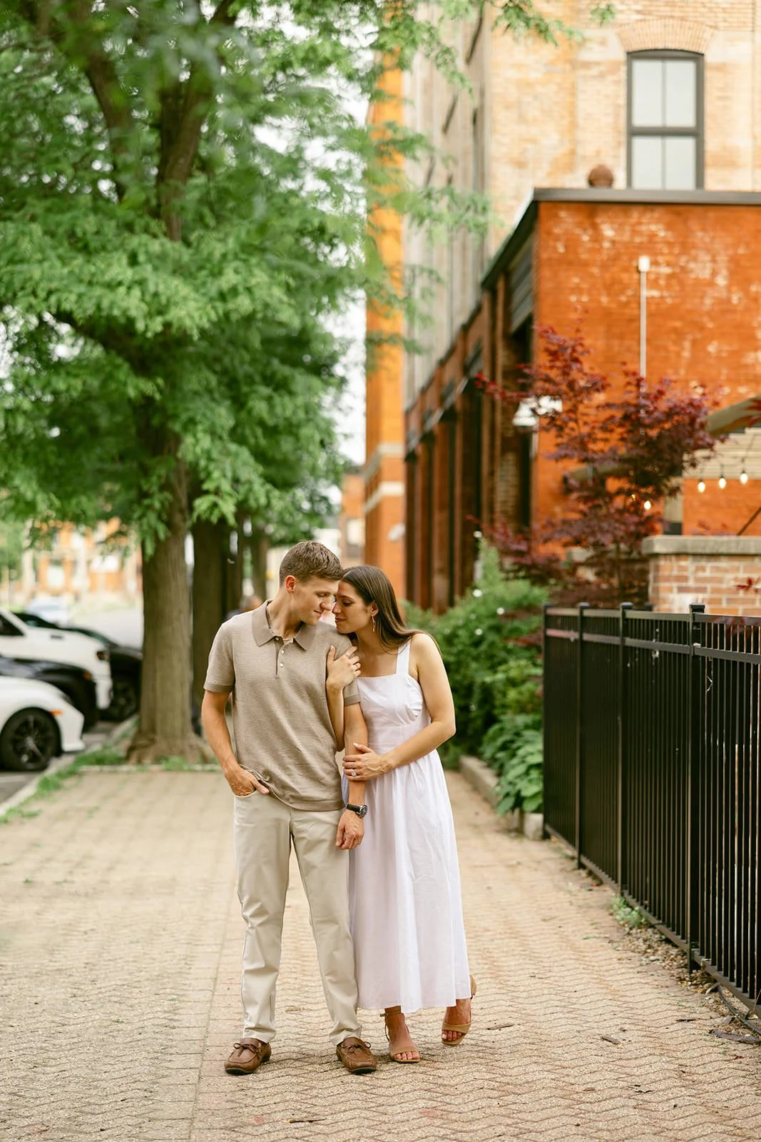 Engaged couple walking arm-in-arm along a brick sidewalk in downtown Utica, NY, surrounded by leafy trees and historic brick buildings. She wears a white midi dress and he wears a neutral polo with light pants.