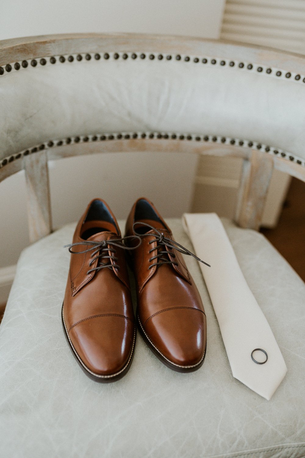 The grooms dress shoes, tie and ring sitting on the chair.