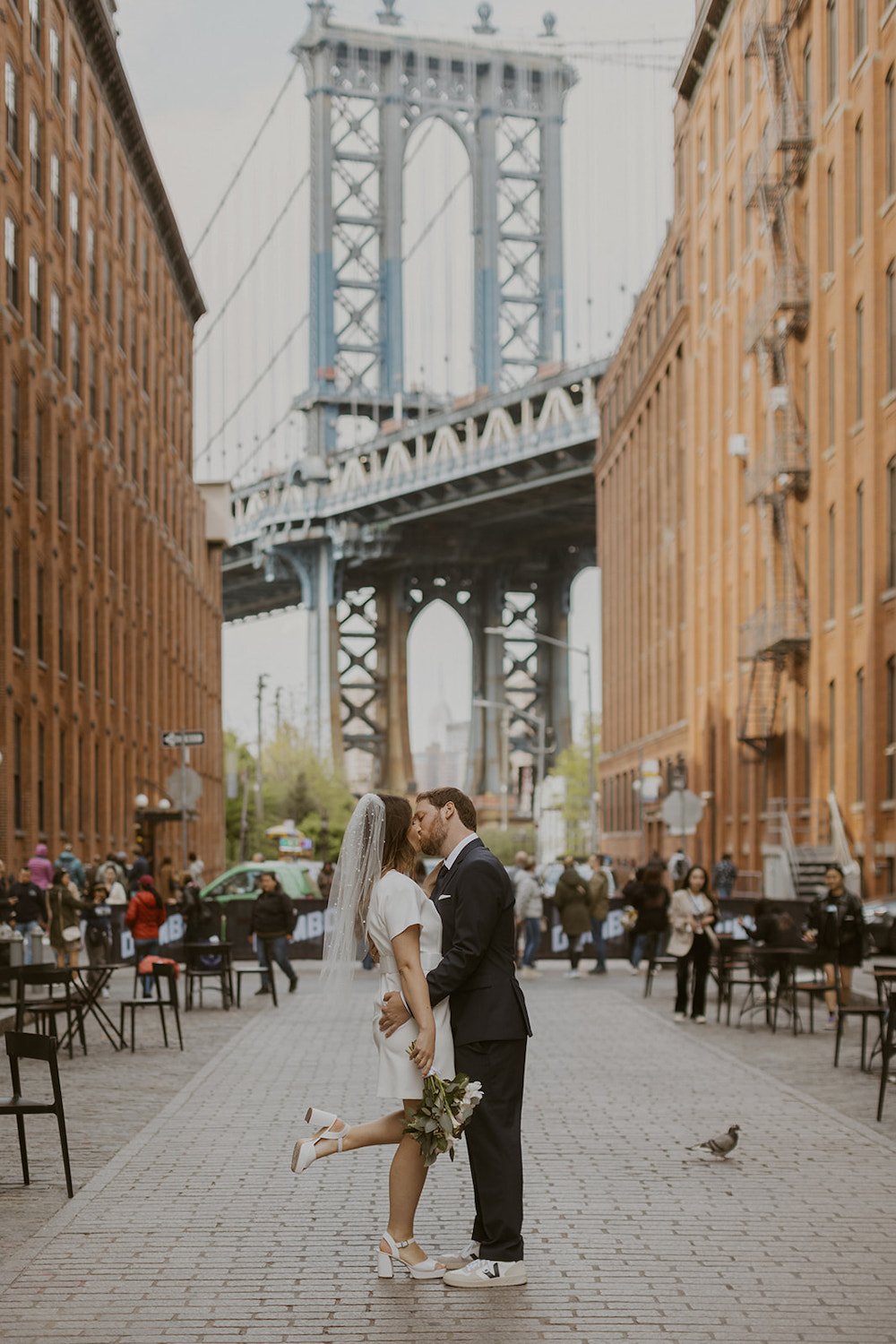 Husband kisses his new wife in the DUMBO district with the Brooklyn Bridge and historic brick building background.