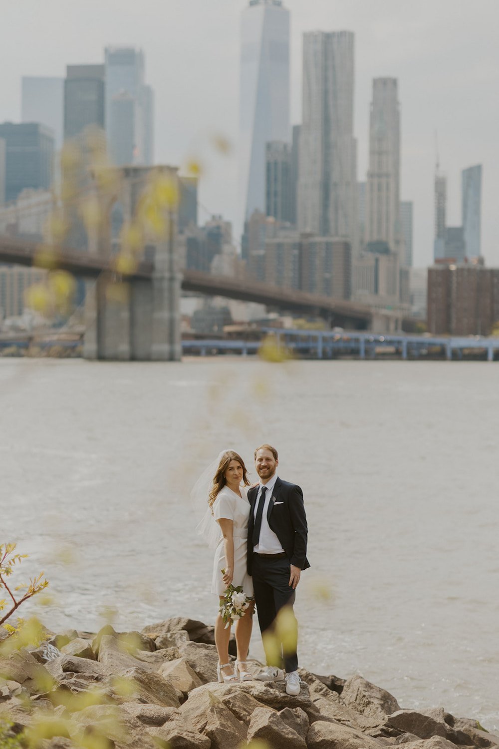 Couple standing on the rocks near the water with the Brooklyn Bridge and City scape in the background.
