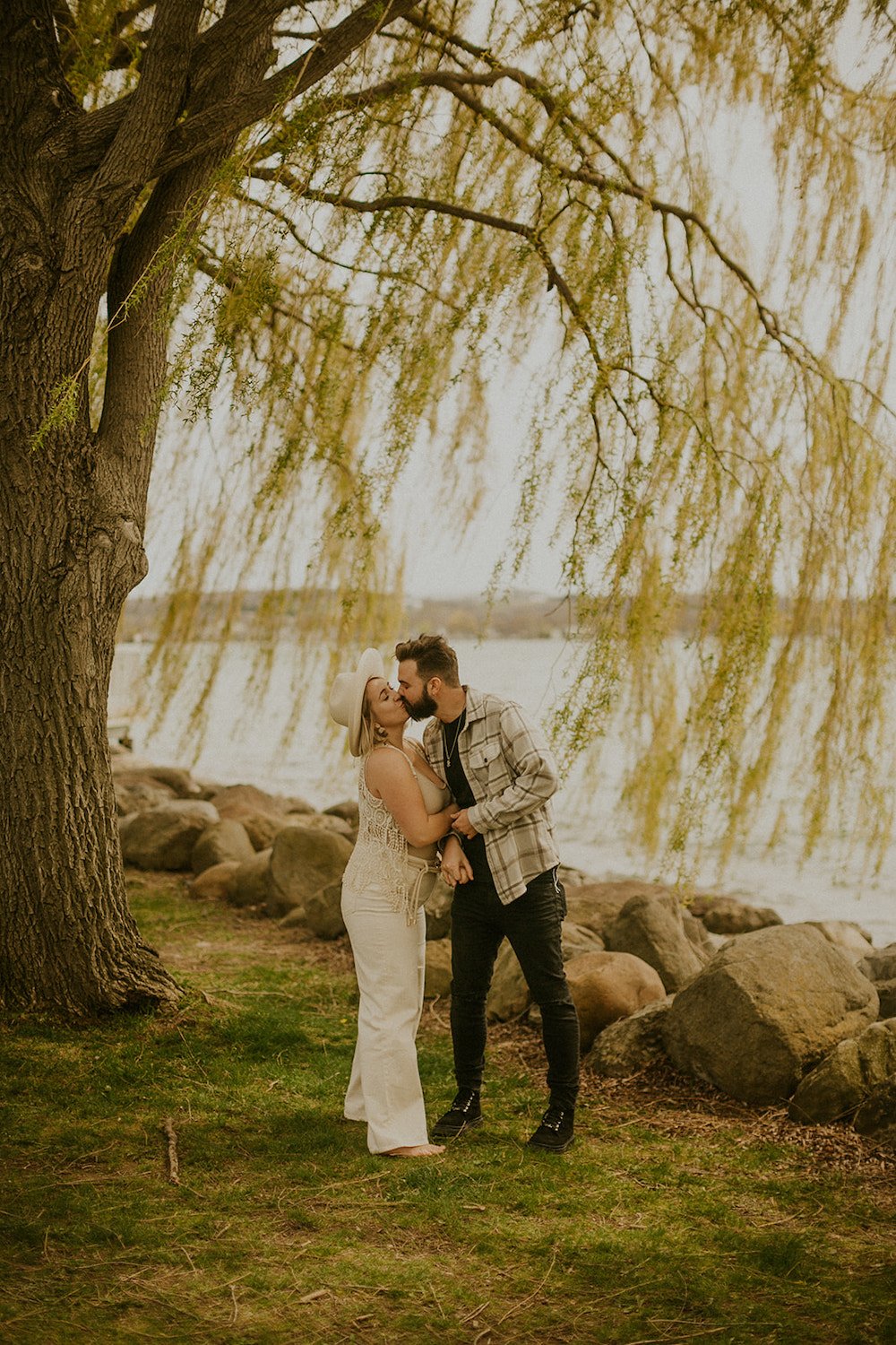 Couple stealing a kiss under the willow tree infront of the lake.