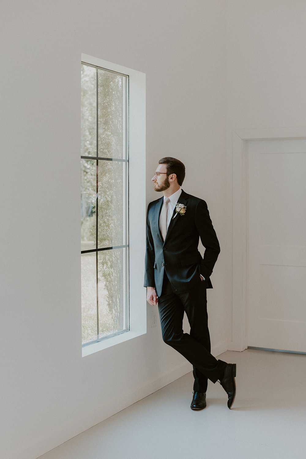 Groom leaning against the wall inside the venue looking out the window dressed in his black tux.