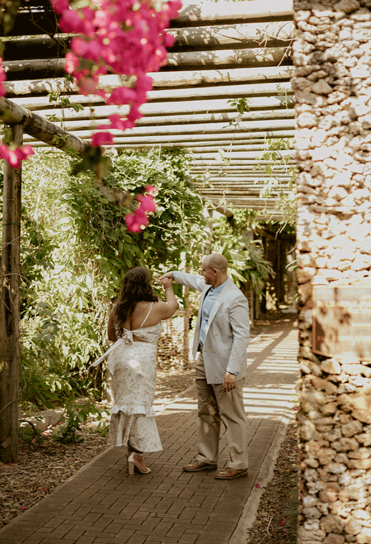 Alex twirls his bride under the archway with the wind blowing the pink and green florals around them.