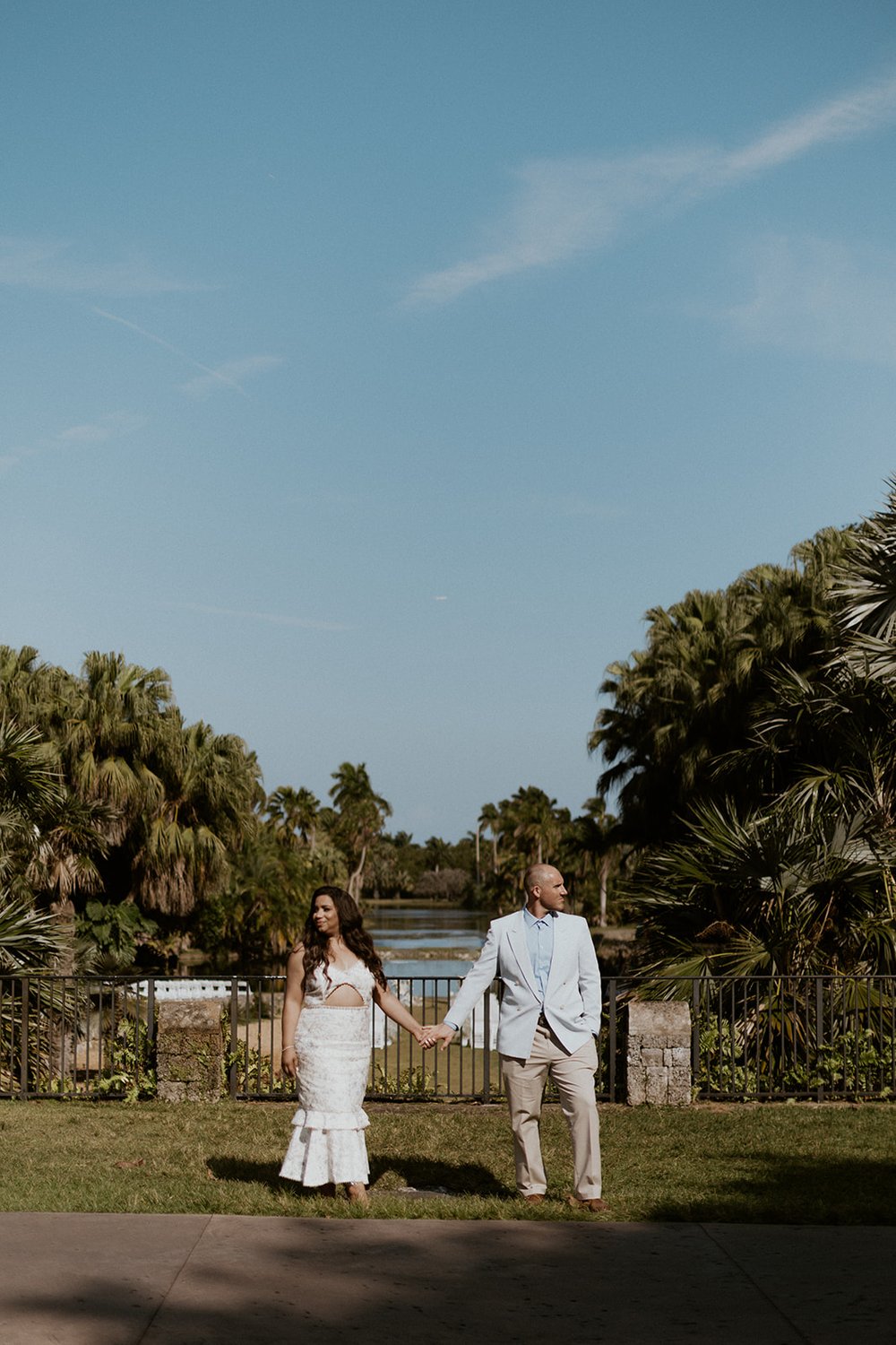 Couple stands in front of the water with a backdrop of lush green trees and a pond. 