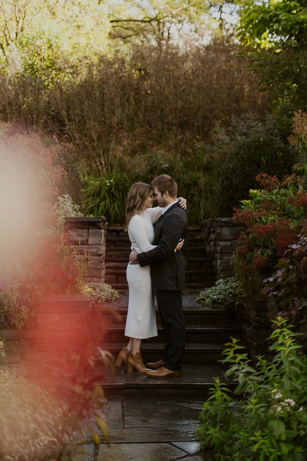  The couple stand on the steps holding one another close wrapped in one another’s love. Greenery surrounds the couple with dark stone stopes.  