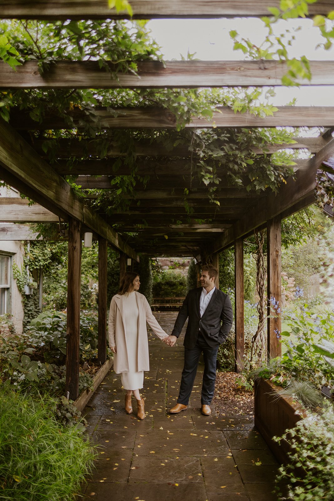  Couple walks holding hands under greenery covered arbor. 