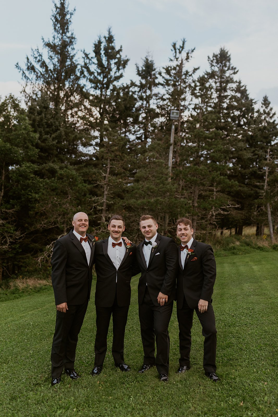 Groomsmen stand together smiling with lush green trees as their background. 