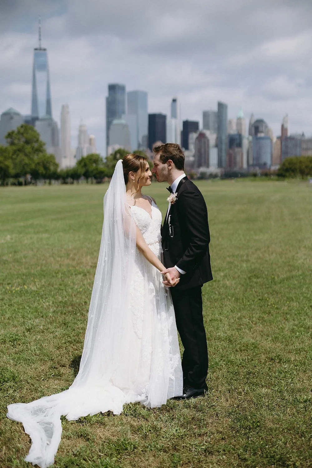 Bride and groom stand in field facing one another. The city sits in the background. 