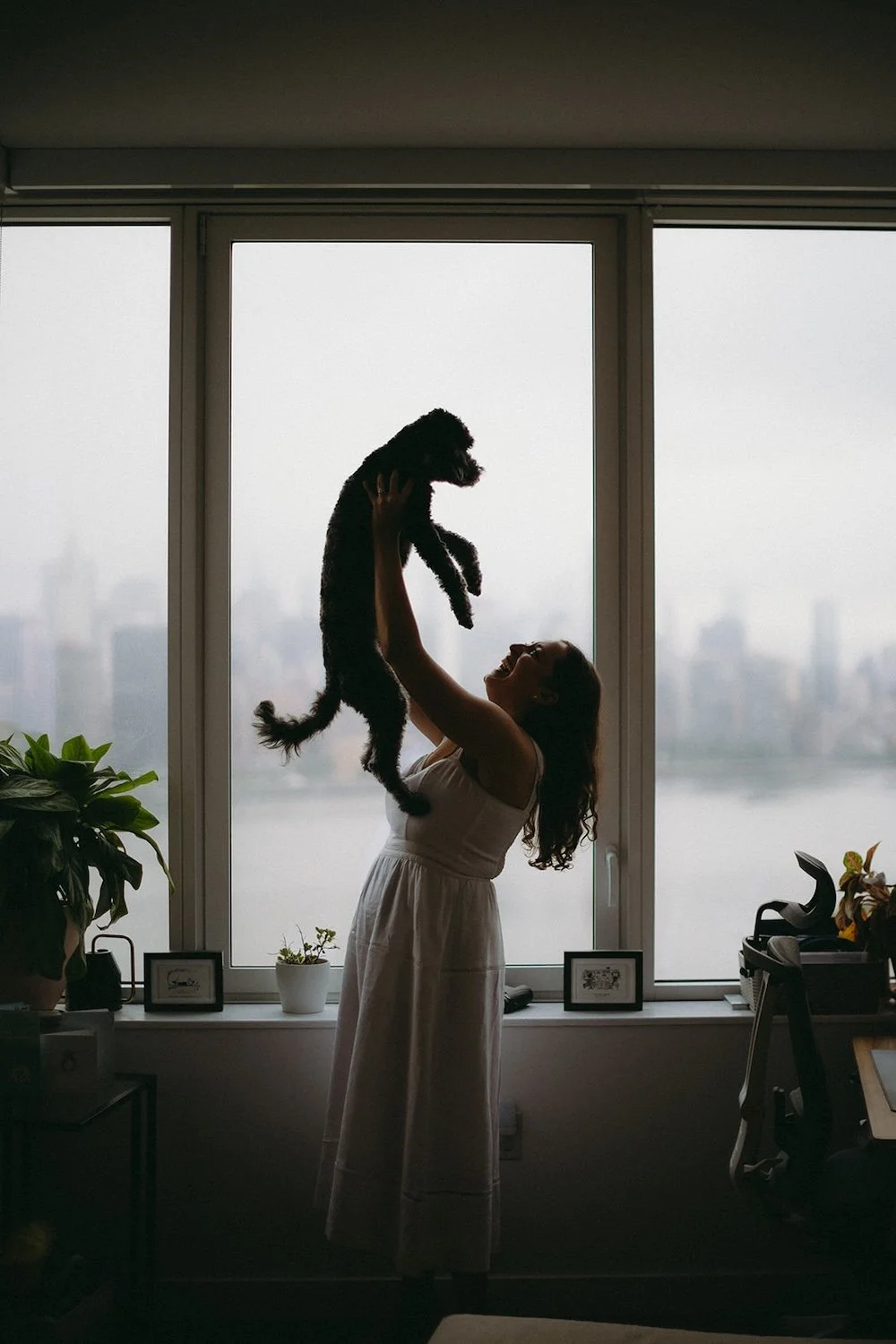 The future bride lifts her dog up in the air while standing infront of the window. 