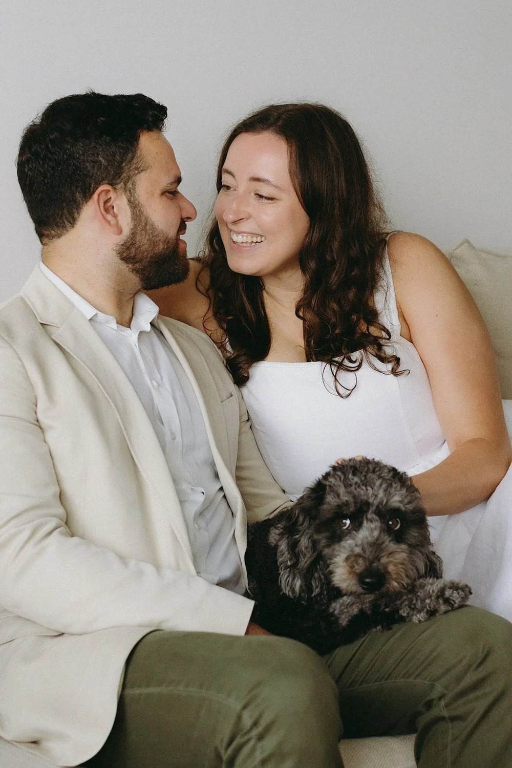 The couple sit on the couch sharing a smile while snuggling their pup. 