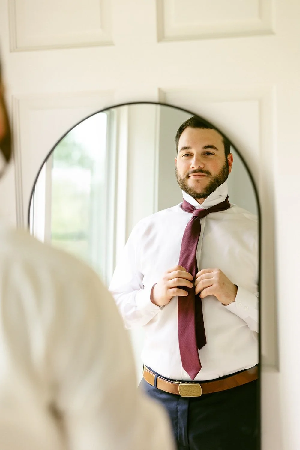 The groom stands in the mirror while tying his tie. 
