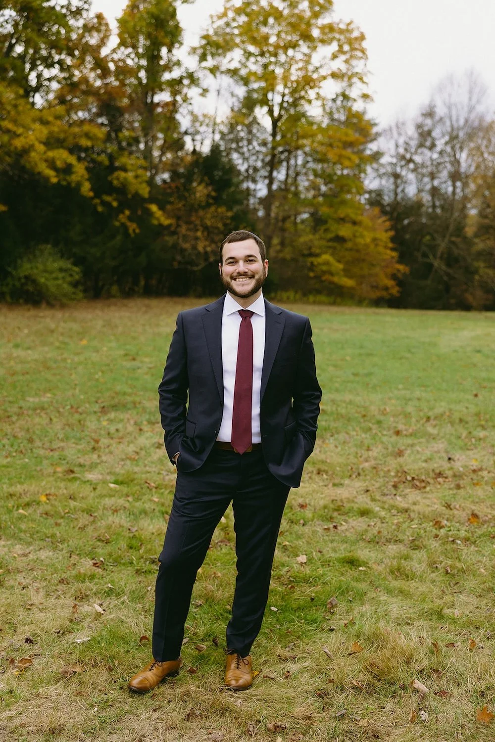 The groom stands in the field with the fall foliage decorated behind him. 