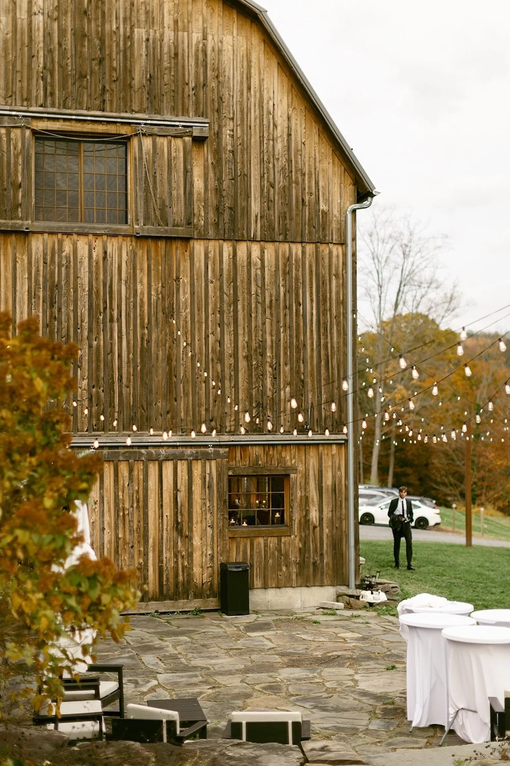 View of the outdoor patio of Homestead Blessings Barn. Lights hanging over the patio with outdoor tables. 