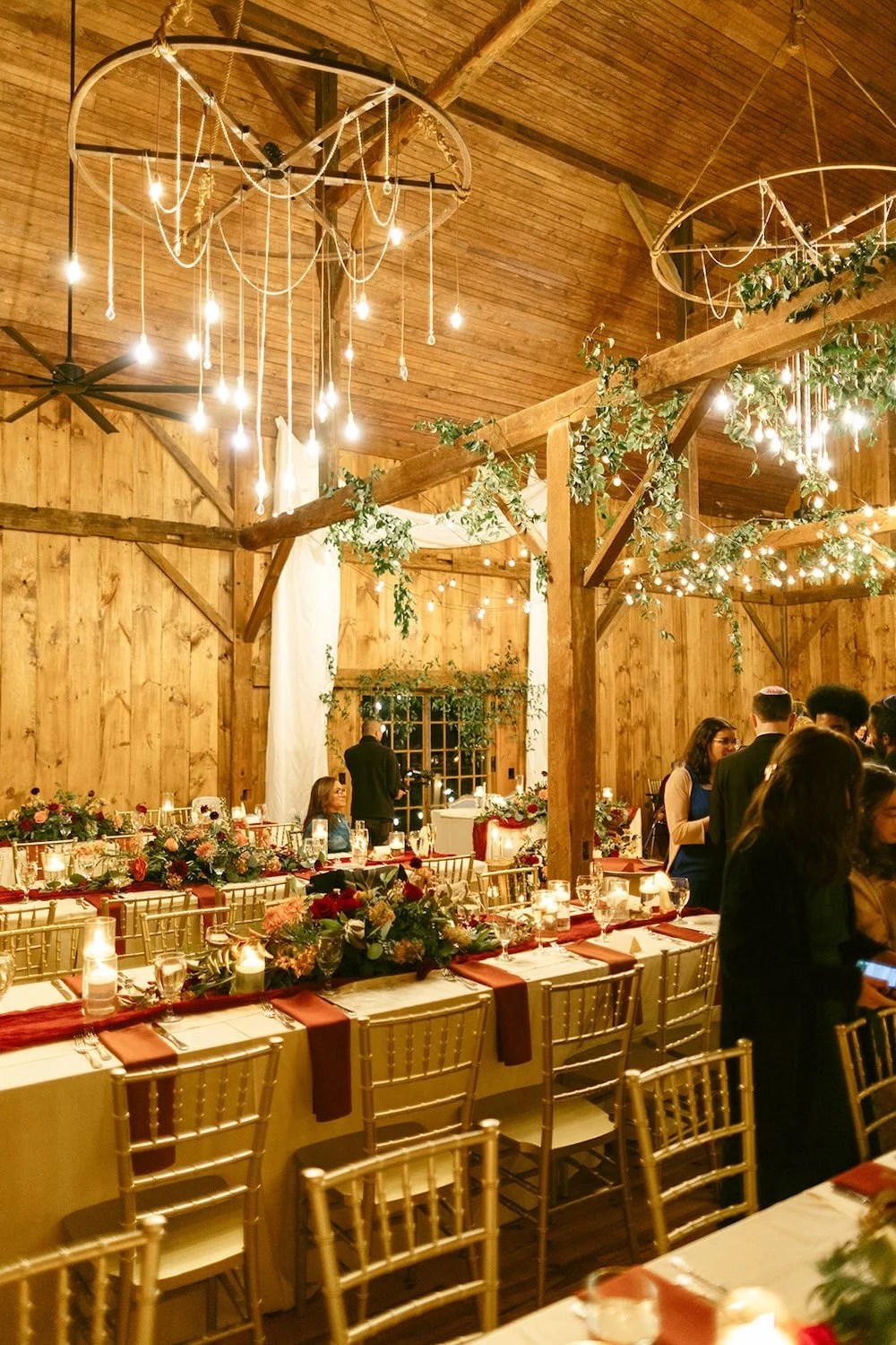 The reception tables decorated with golds and maroons. Hanging lights brighten the room and bring out the oak wood of the barn. 