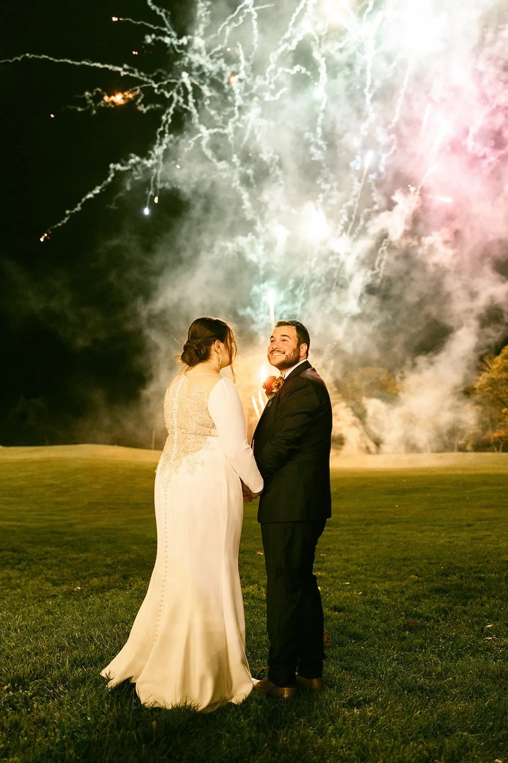 The groom looks back during the fireworks with a bright smile while he holds his bride close. 