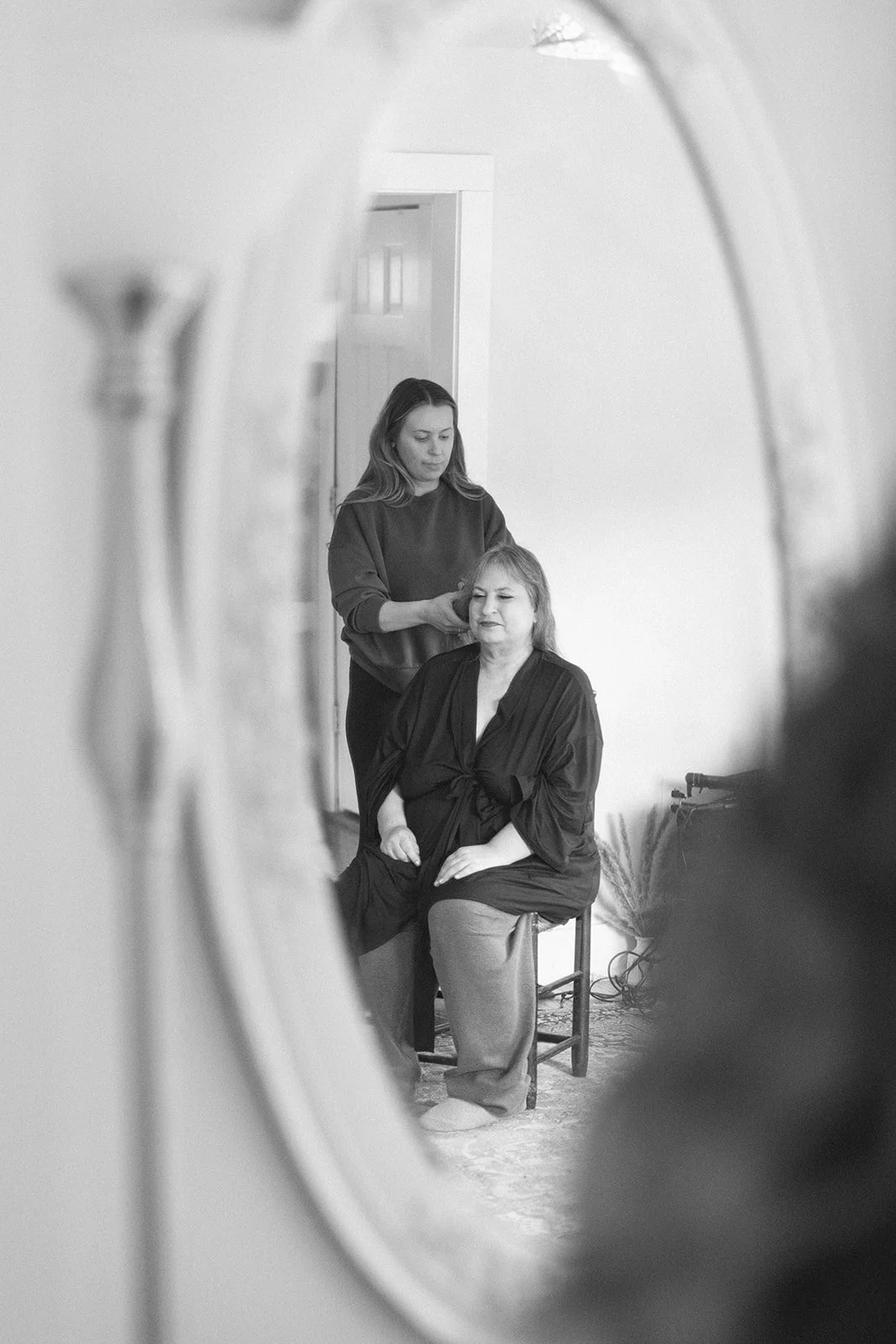 Mother of the bride sits while getting her hair done. View of her through a mirror. 
