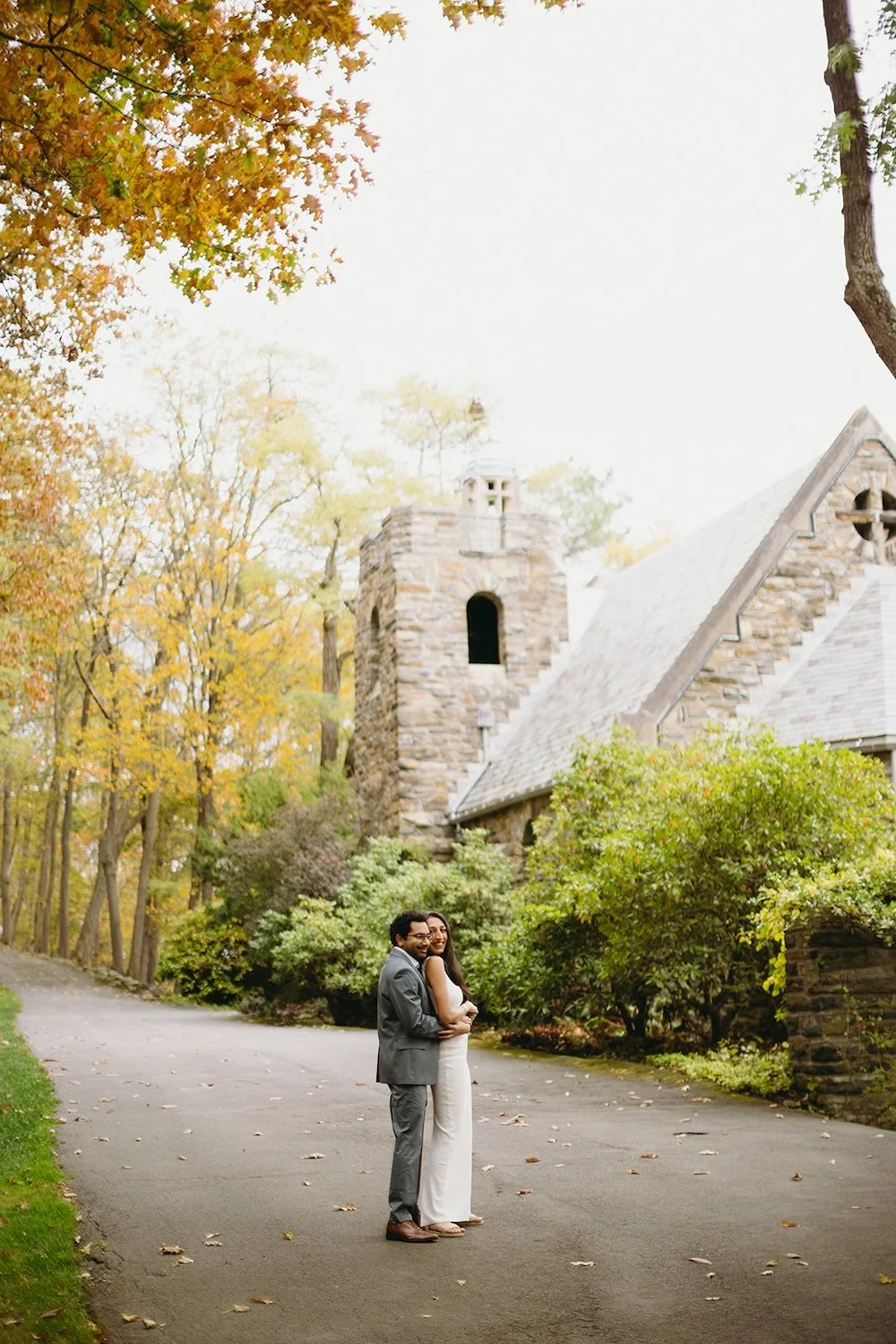 The couple stand close together with the church and the trees surrounding them.