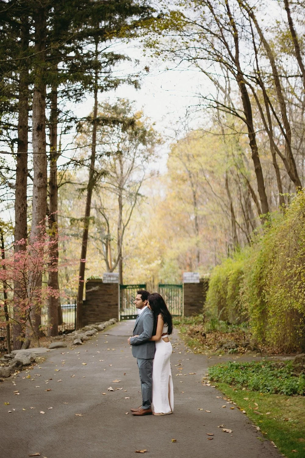 The couple stand infront of the fence entry way of the church as the future bride holds him close. 