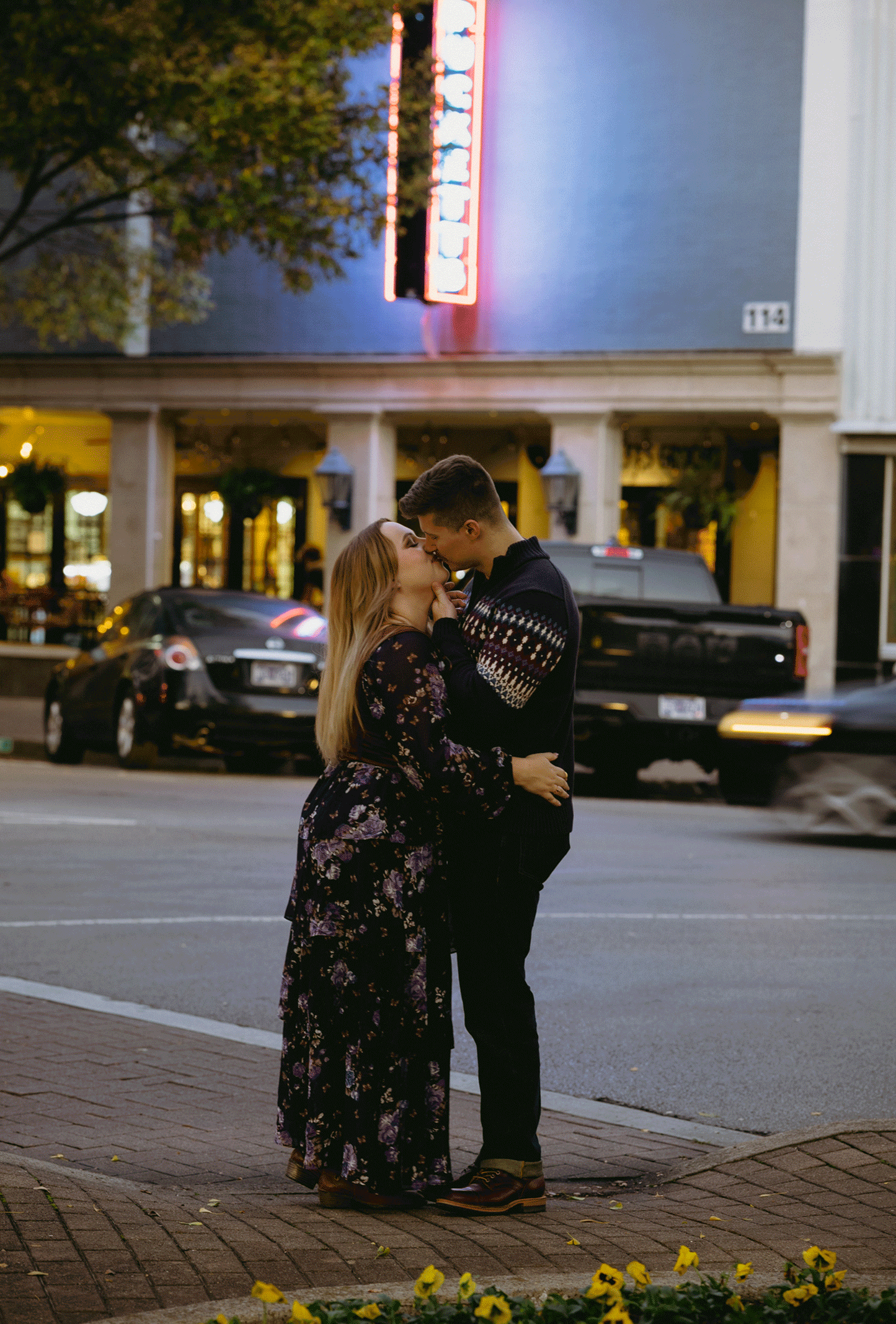 The future groom kisses his bride to be at the corner of downtown with cars racing by behind them.
