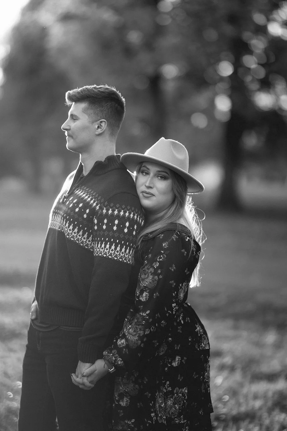 Black and white photo of the couple male stands looking out with female resting her head on his back while holding his hands. 